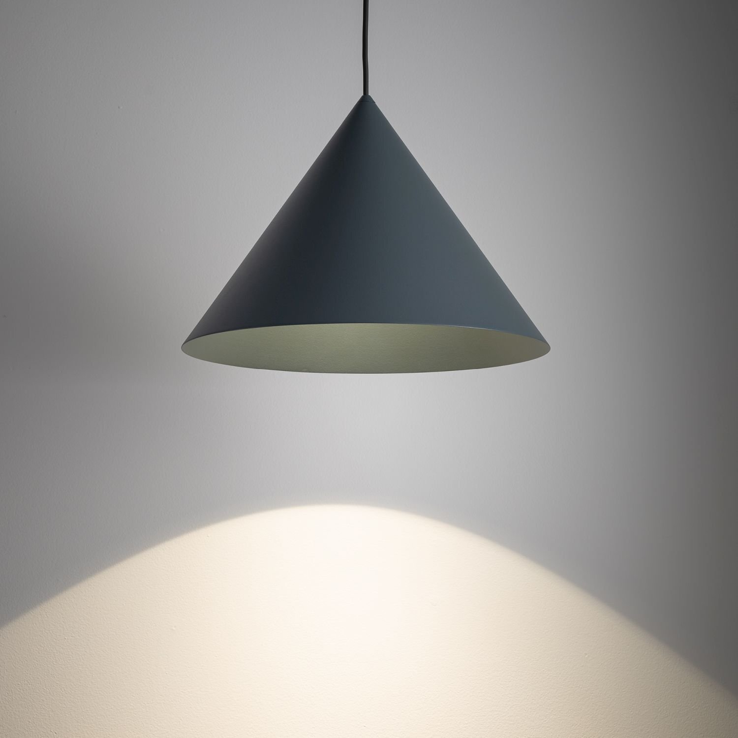 Na obrazie widoczna jest lampa wisząca o stożkowym kształcie, emitująca światło na ścianę. Lampa ma ciemny kolor zewnętrzny i jasne wnętrze.