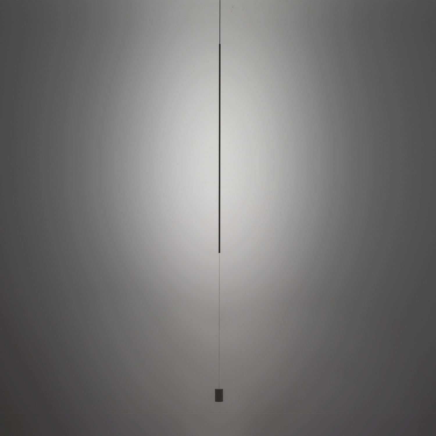 Na obrazie widoczna jest smukła, pionowa lampa wisząca LED z czarnym wykończeniem, zawieszona na cienkim przewodzie. Tło jest jednolite i szare, co podkreśla minimalistyczny design lampy.