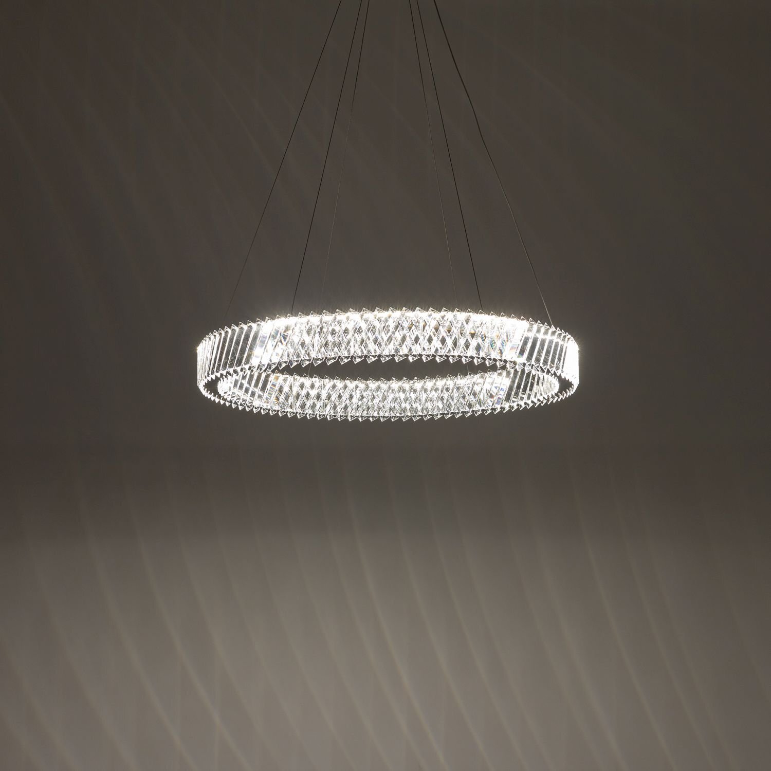 Na obrazie widoczna jest nowoczesna lampa wisząca w kształcie okręgu, emitująca jasne światło. Lampa zawieszona jest na cienkich przewodach, a jej powierzchnia ma strukturę przypominającą kryształki.