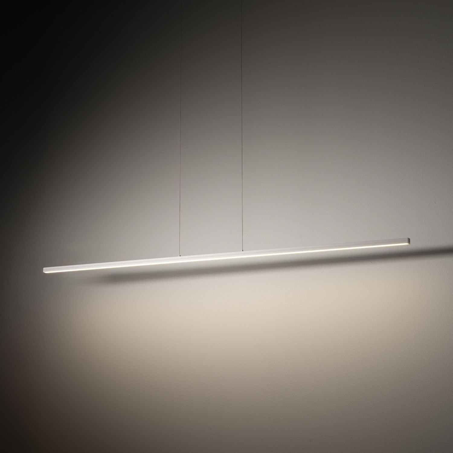 Na obrazie widoczna jest nowoczesna lampa wisząca LED o prostym, liniowym kształcie, zawieszona na dwóch cienkich przewodach. Światło lampy tworzy delikatny cień na ścianie.