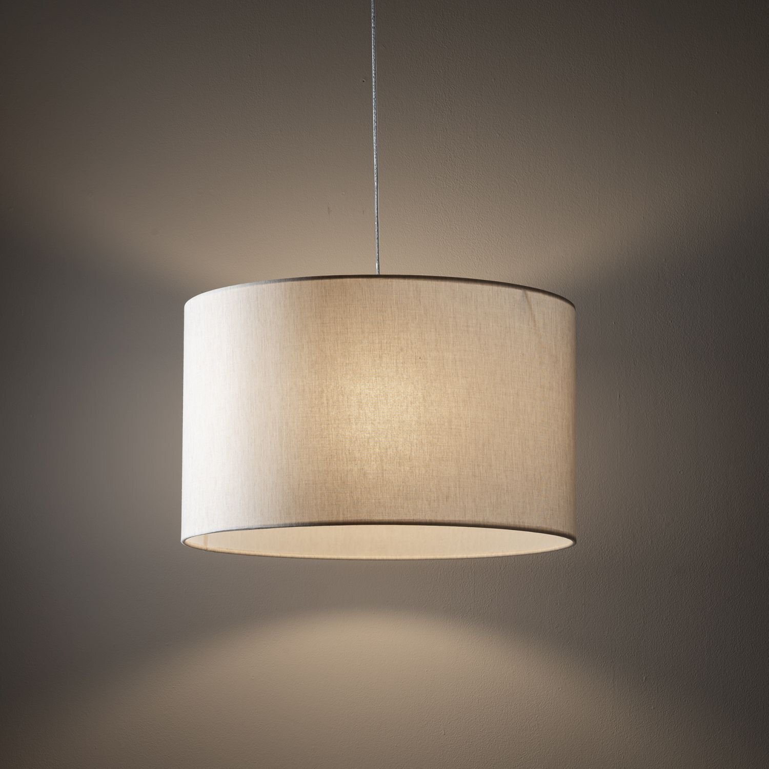 Na obrazie widoczna jest lampa wisząca z cylindrycznym, jasnym abażurem, zawieszona na cienkim przewodzie. Światło delikatnie oświetla ścianę w tle.