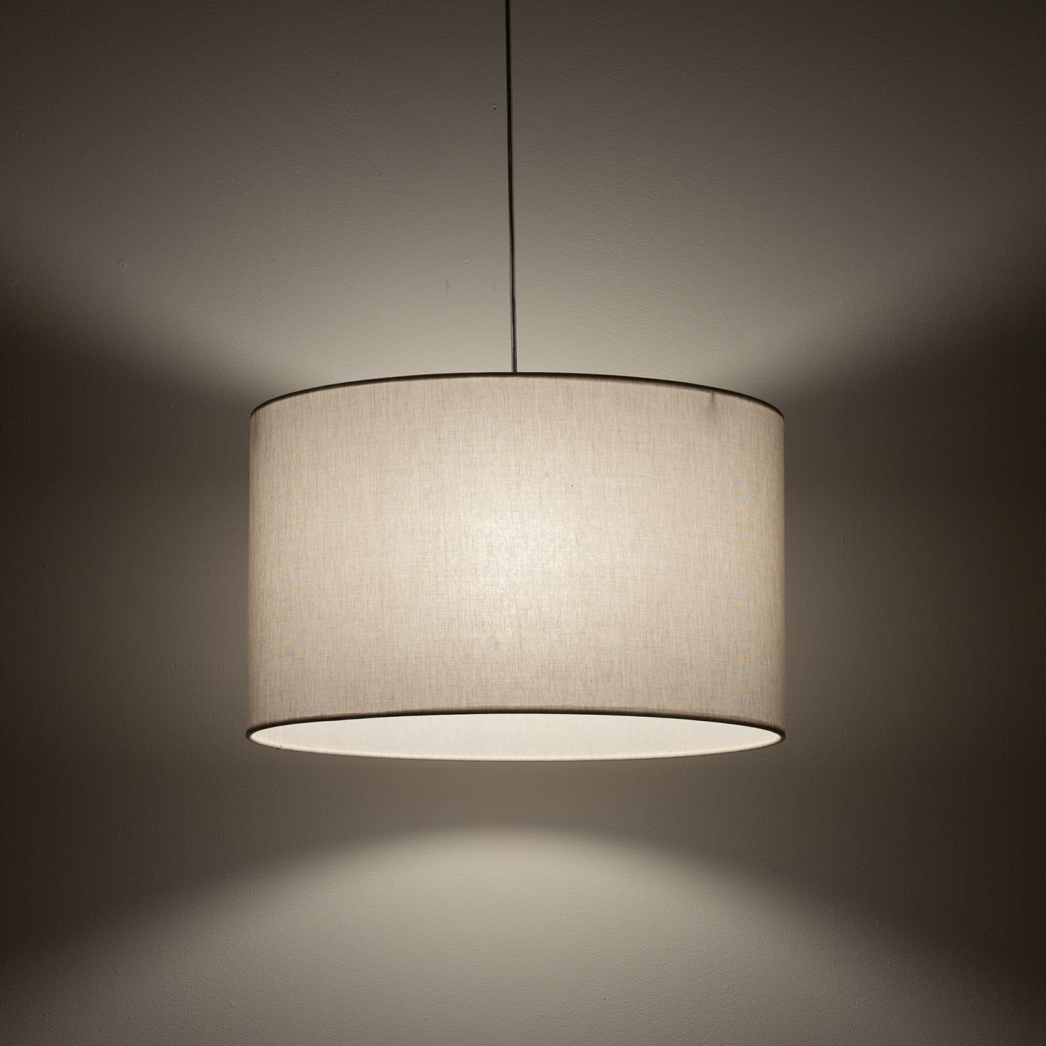 Na obrazie widoczna jest wisząca lampa z jasnym, cylindrycznym abażurem, emitująca ciepłe światło. Lampa zawieszona jest na cienkim przewodzie, a jej światło tworzy delikatne cienie na ścianie.