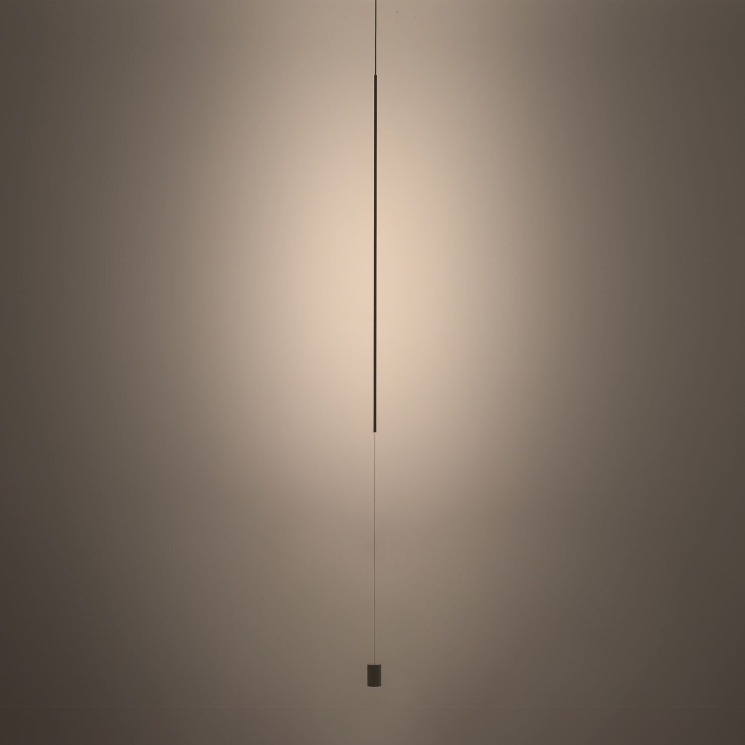 Na obrazie widoczna jest smukła, pionowa lampa wisząca LED, emitująca delikatne światło na ścianę. Lampa jest zawieszona na cienkim przewodzie, a jej dolna część zakończona jest małym, cylindrycznym elementem.