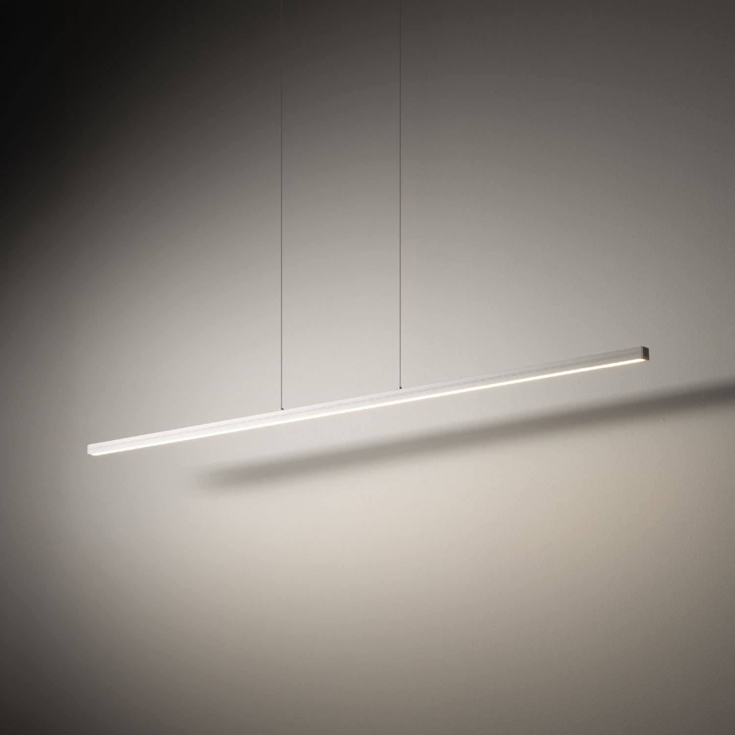 Na obrazie widoczna jest nowoczesna lampa wisząca o prostym, liniowym kształcie, emitująca jasne światło. Lampa zawieszona jest na dwóch cienkich przewodach na tle jednolitej, jasnej ściany.