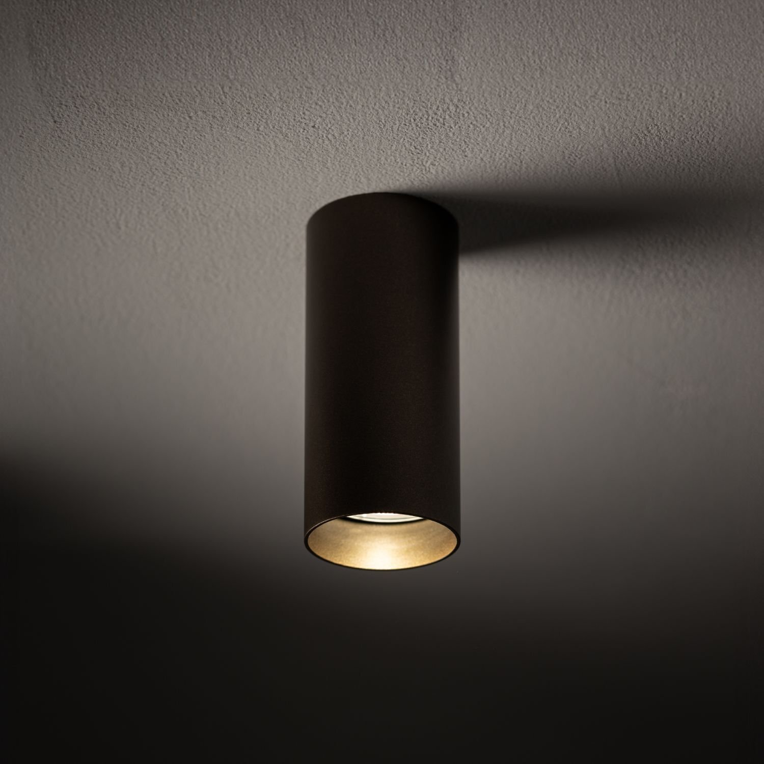 Cylindryczna lampa sufitowa w ciemnym kolorze, emitująca światło skierowane w dół. Lampa jest zamontowana na gładkim suficie.