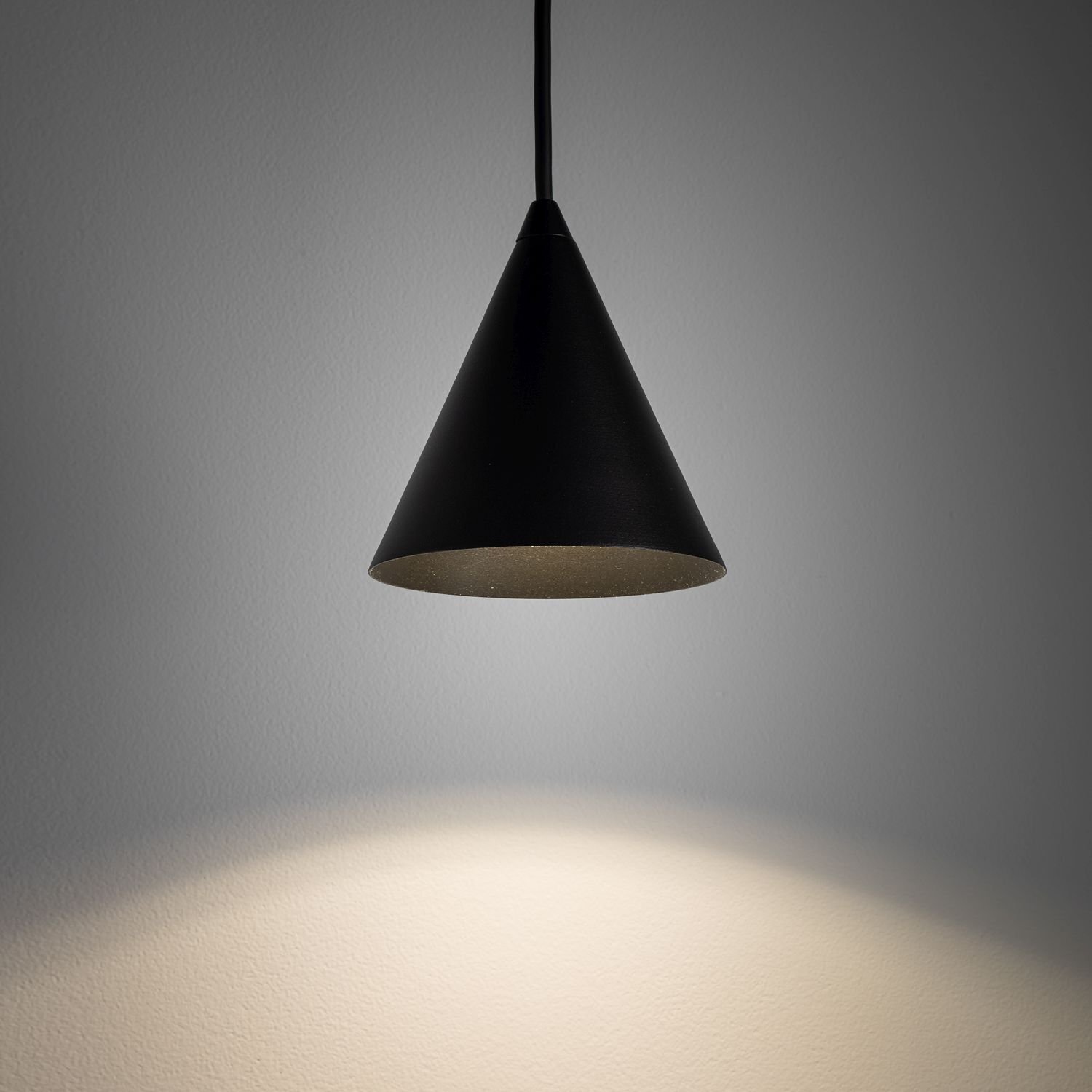 Czarna, stożkowa lampa wisząca oświetla ścianę miękkim, białym światłem.