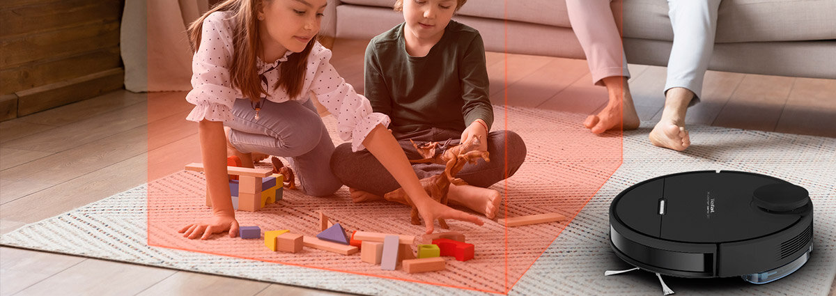 Dwoje dzieci bawi się klockami na dywanie, obok znajduje się czarny robot sprzątający. Robot jest włączony i widoczny jest czerwony obszar wokół dzieci, sugerujący strefę bezpieczeństwa.