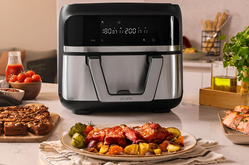 Na blacie kuchennym stoi frytkownica beztłuszczowa z wyświetlaczem pokazującym temperatury 180°C i 200°C. Przed urządzeniem znajduje się talerz z pieczonymi warzywami i mięsem.