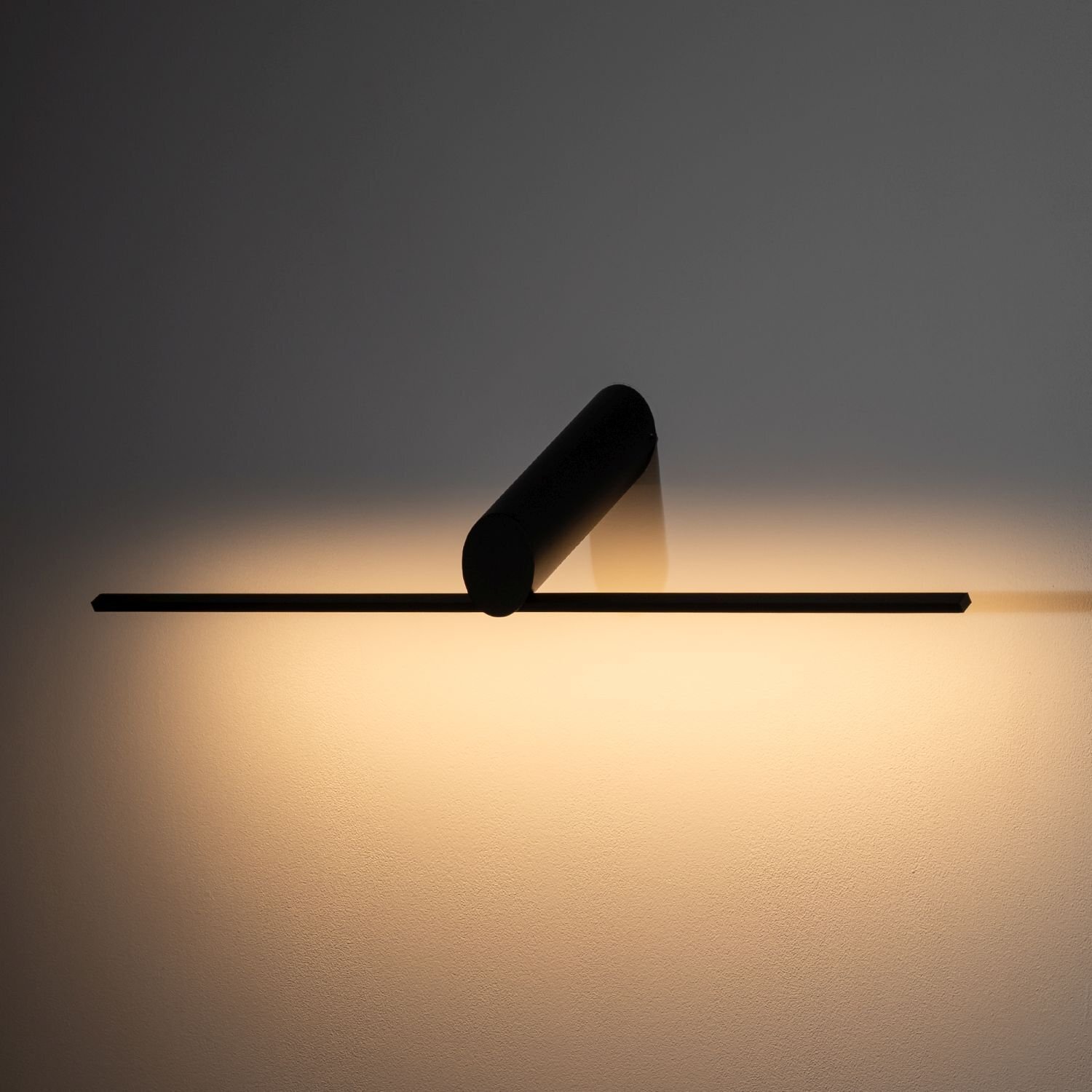 Na obrazie widoczna jest nowoczesna lampa LED zamontowana na ścianie, emitująca ciepłe światło. Lampa ma prosty, cylindryczny kształt i jest w kolorze czarnym.