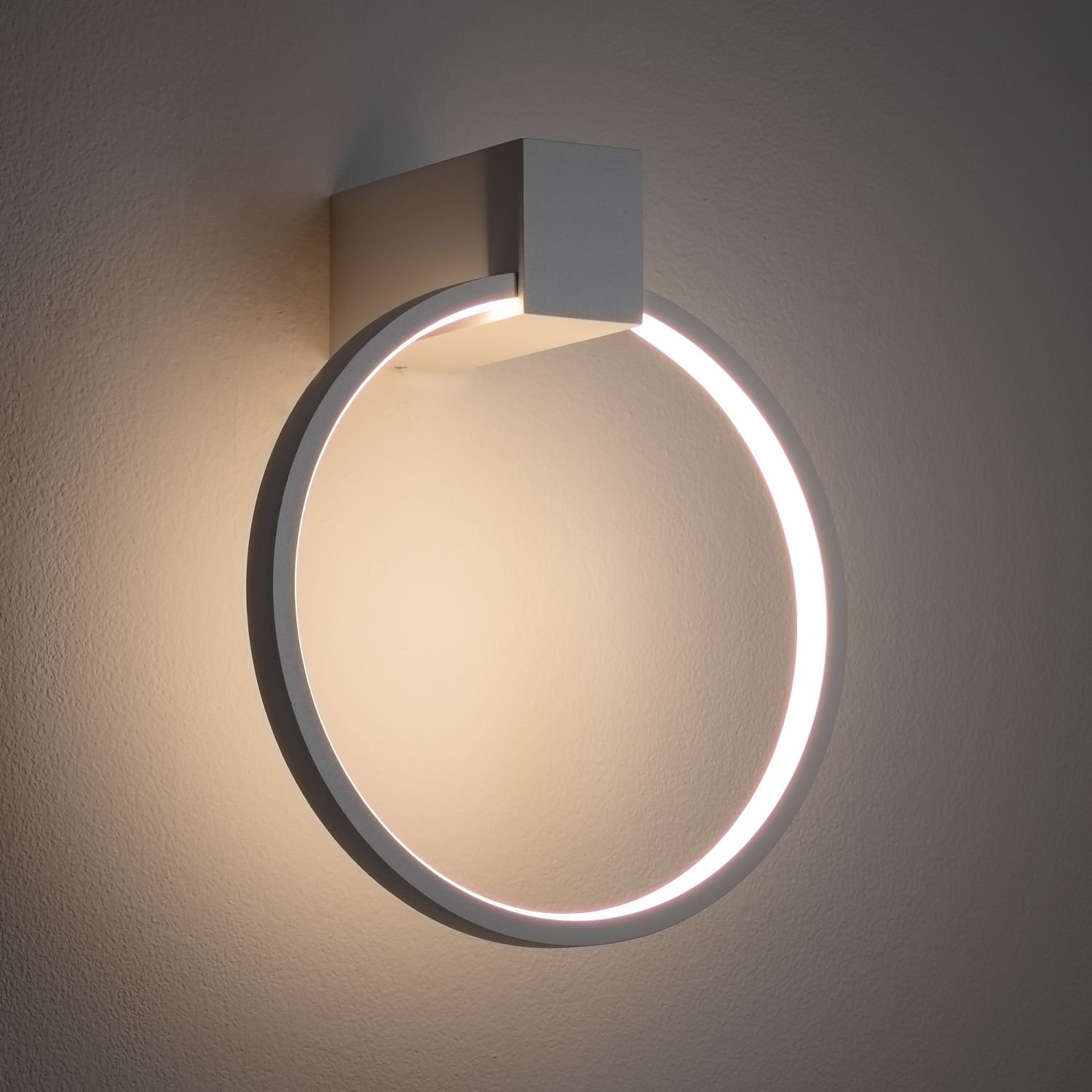 Na obrazie widoczny jest nowoczesny kinkiet LED w kształcie okręgu, emitujący jasne światło. Lampa jest zamontowana na ścianie i ma minimalistyczny design.