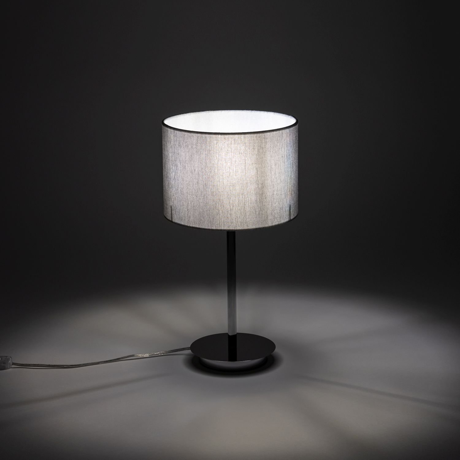 Na obrazie widoczna jest elegancka lampa stołowa z cylindrycznym, jasnym abażurem i ciemną podstawą. Lampa stoi na ciemnym tle, a jej kabel jest widoczny na powierzchni.