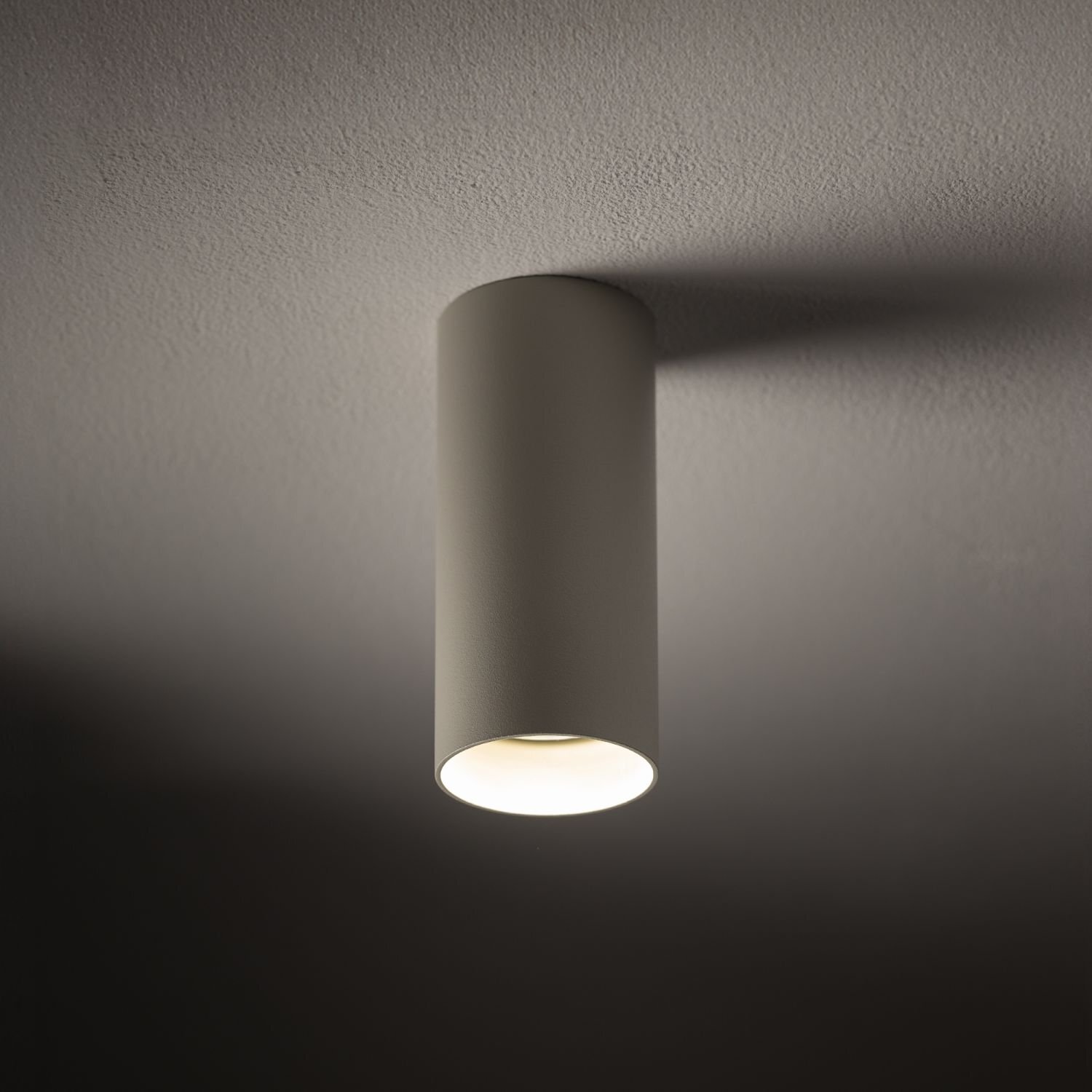 Cylindryczna lampa natynkowa w kolorze szarym zamontowana na suficie, emitująca światło w dół. Tło jest jednolite i ciemne.