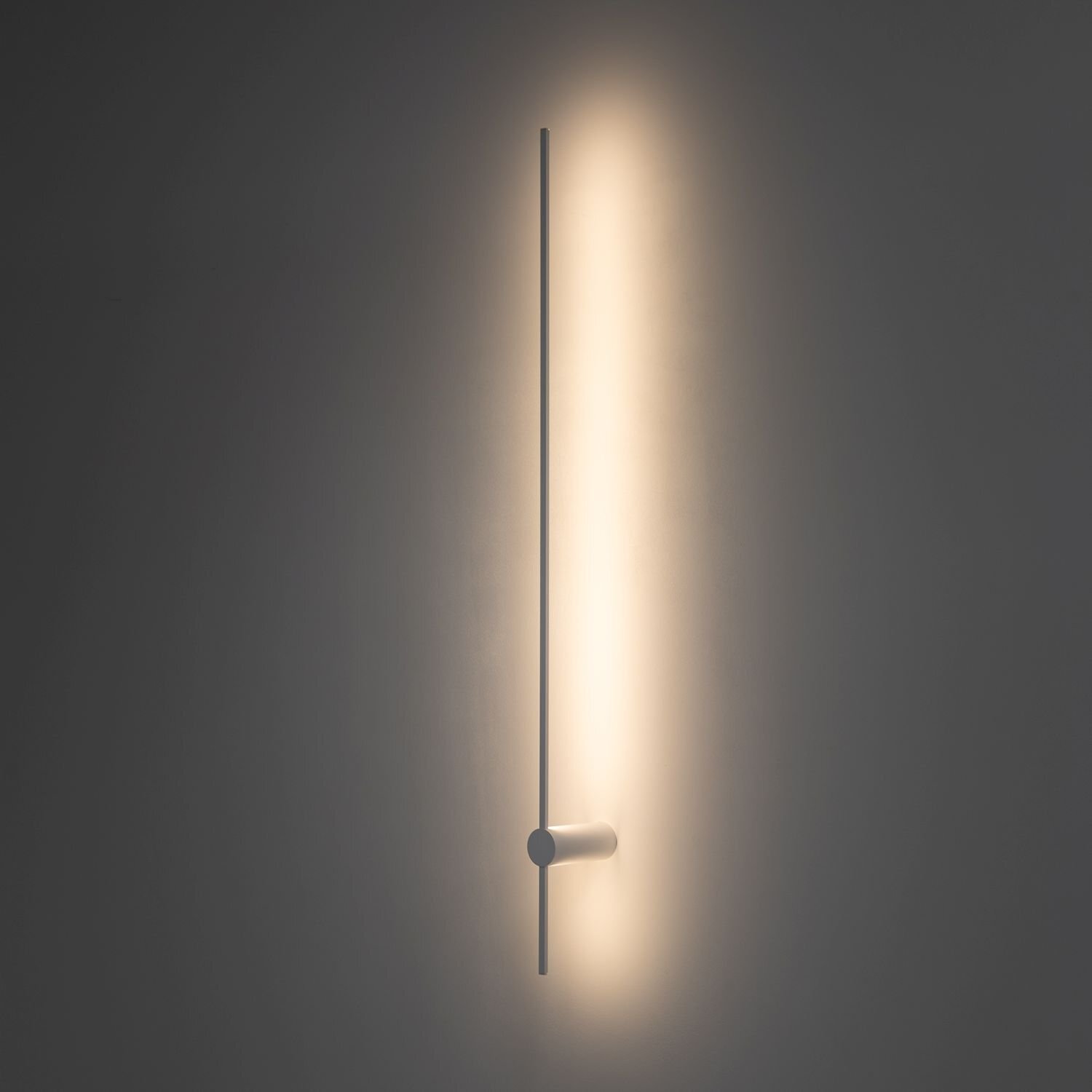 Na obrazie widoczna jest nowoczesna lampa ścienna emitująca jasne, rozproszone światło na ciemnym tle. Lampa ma prosty, pionowy kształt z poziomym elementem na dole.