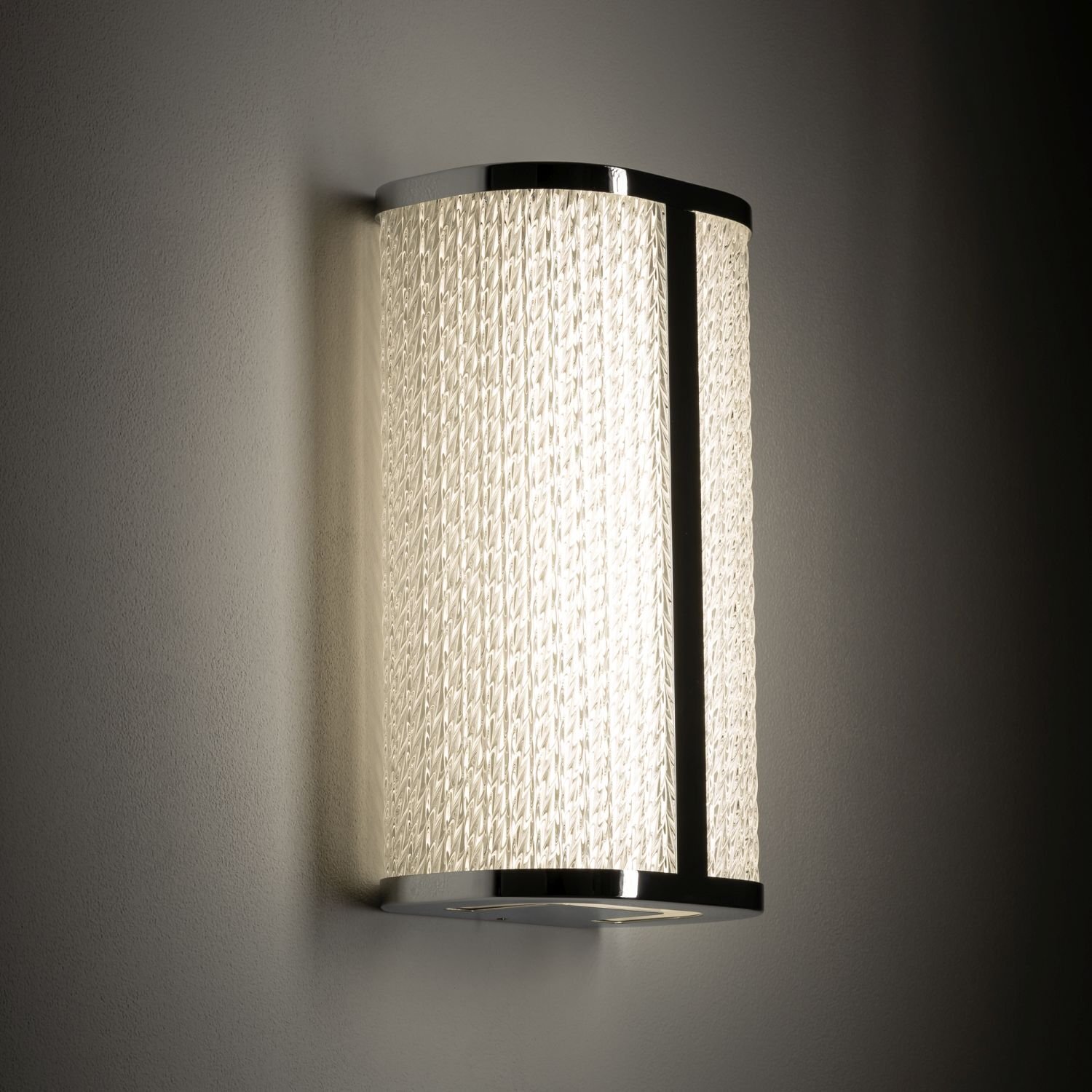 Na obrazie widoczna jest nowoczesna lampa ścienna z jasnym, teksturowanym kloszem i metalowymi elementami wykończenia. Lampa emituje ciepłe, rozproszone światło.