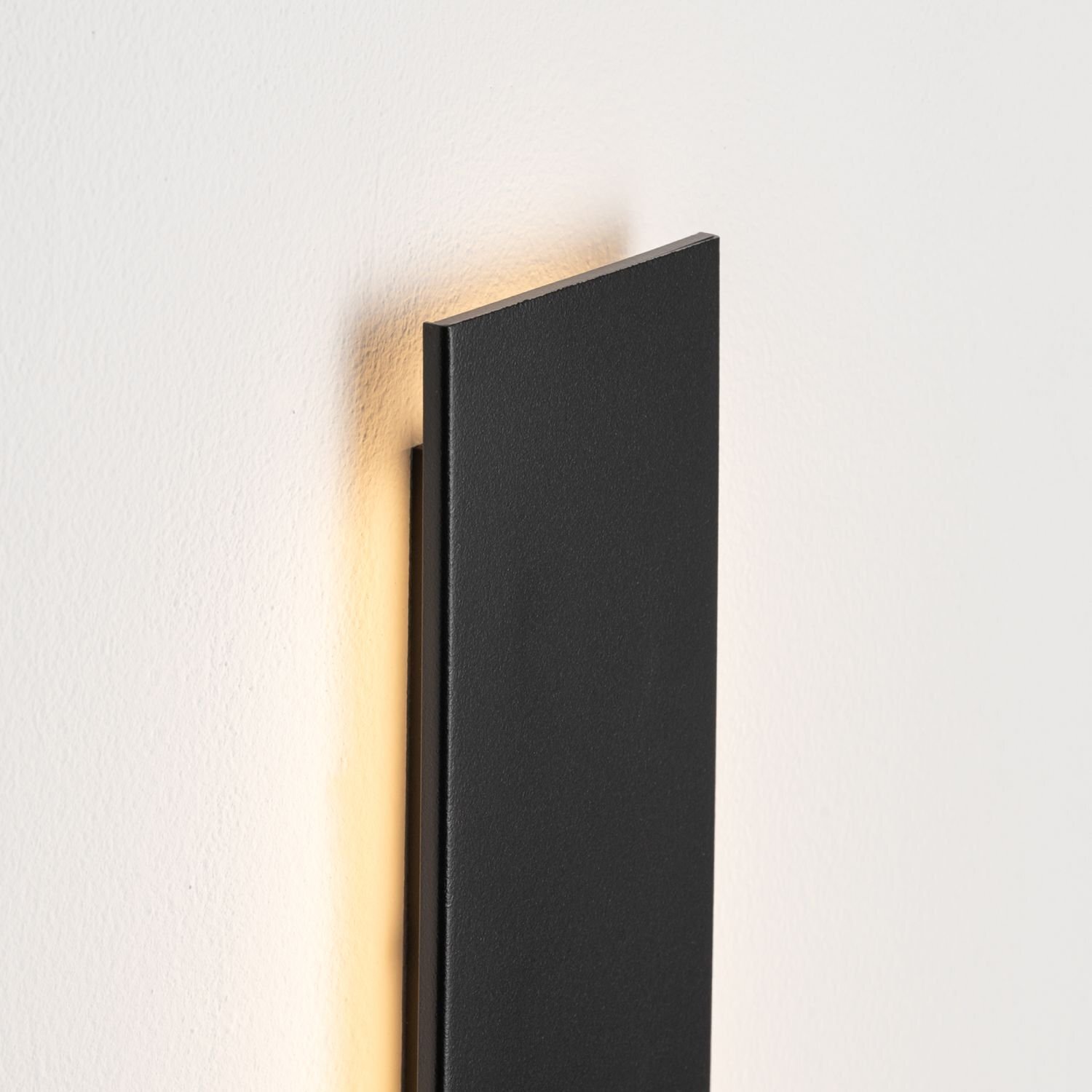 Czarna, prostokątna lampa ścienna LED zamontowana na jasnej ścianie, emitująca delikatne światło z tyłu. Lampa ma nowoczesny, minimalistyczny design.