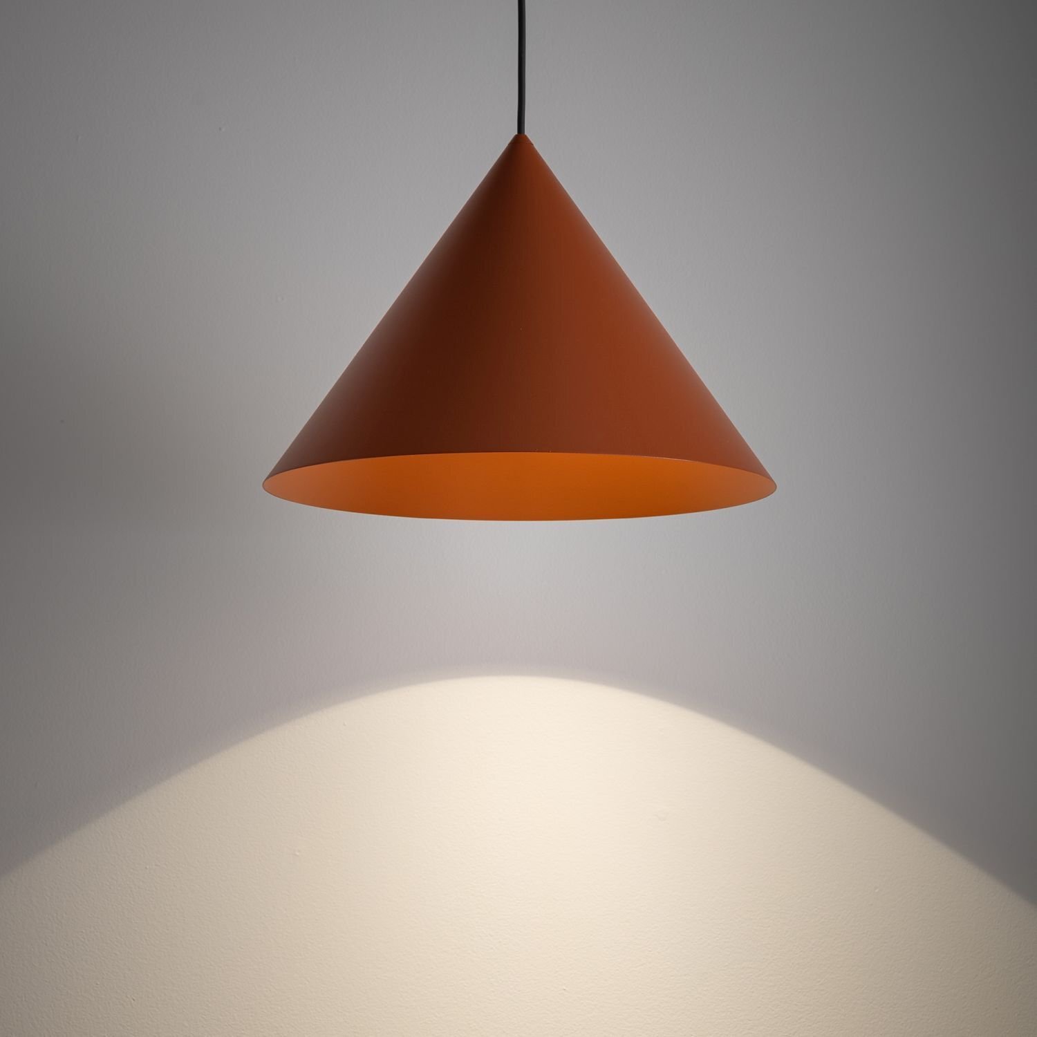 Na obrazie widoczna jest wisząca lampa w kształcie stożka o brązowym kolorze, emitująca światło na ścianę poniżej. Lampa jest zawieszona na cienkim czarnym przewodzie.