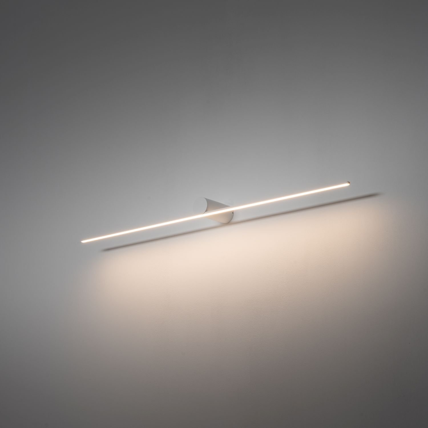 Na obrazie widoczna jest nowoczesna lampa ścienna LED o prostym, liniowym kształcie, emitująca delikatne światło na ścianę. Lampa jest zamontowana na gładkiej, jasnej powierzchni.