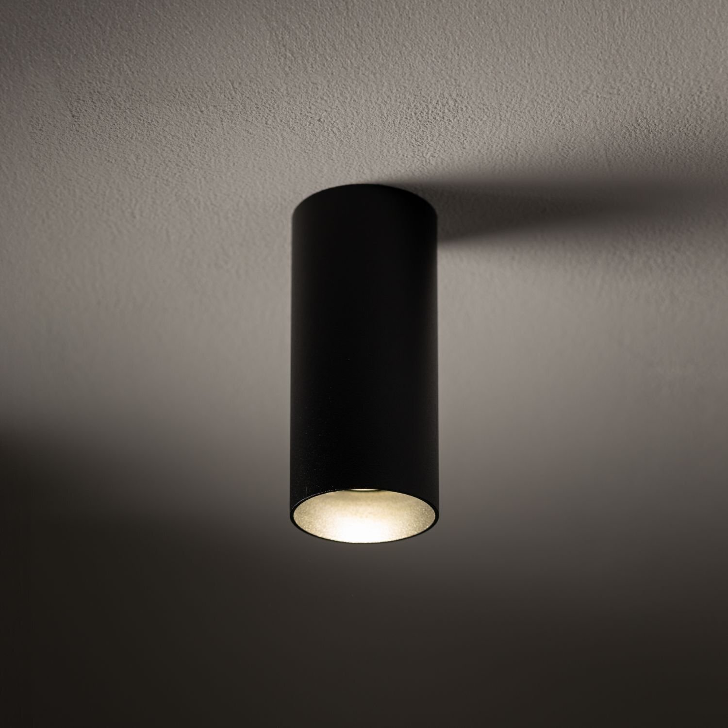 Czarna, cylindryczna lampa natynkowa zamontowana na suficie, emitująca światło w dół. Światło tworzy delikatny cień na suficie.