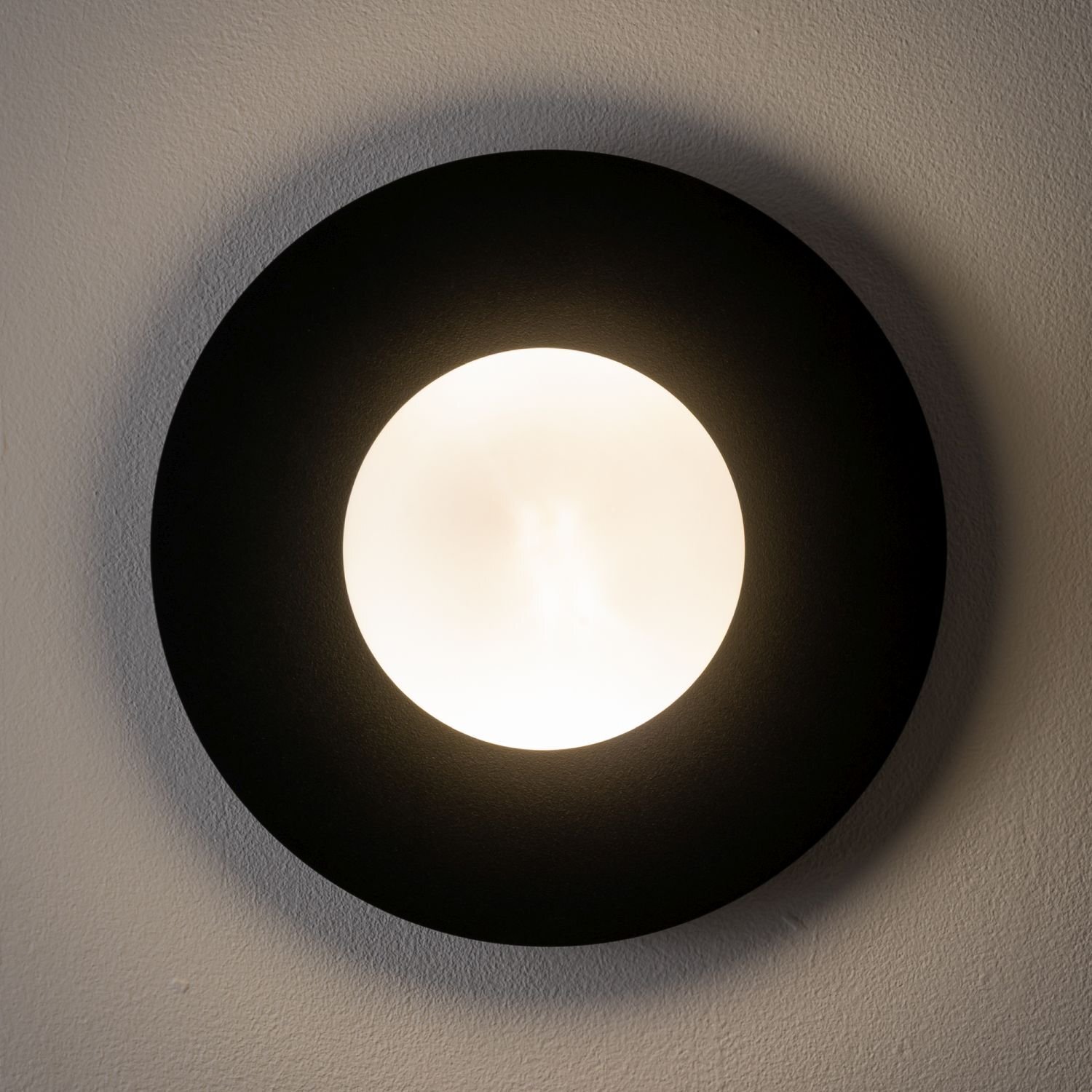 Na obrazie widoczna jest okrągła lampa ścienna z czarną obwódką i jasnym, świecącym środkiem. Lampa jest zamontowana na ścianie.