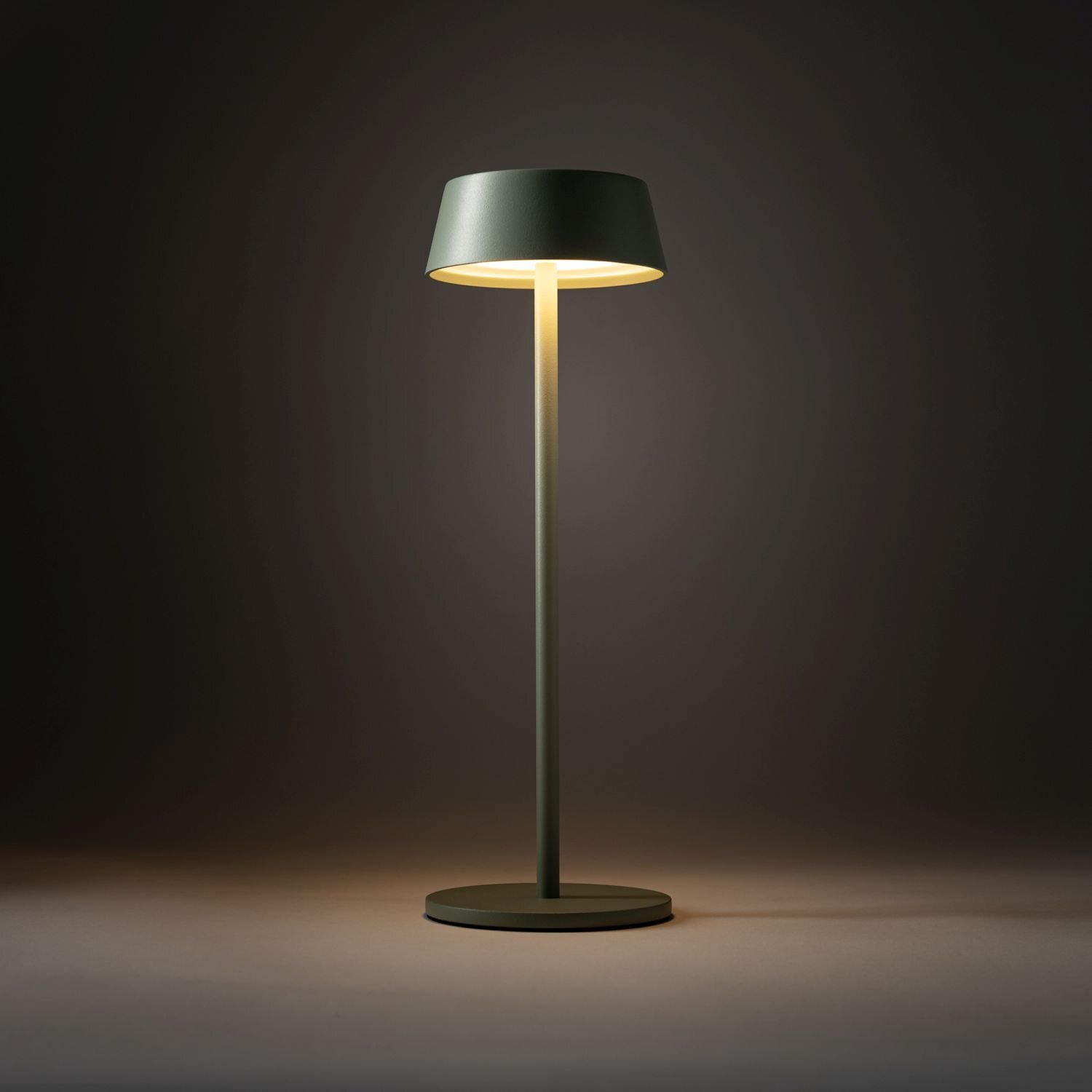 Na obrazie widoczna jest przenośna lampa zewnętrzna LED o prostym, nowoczesnym designie, emitująca ciepłe światło. Lampa ma ciemny klosz i stoi na okrągłej podstawie.