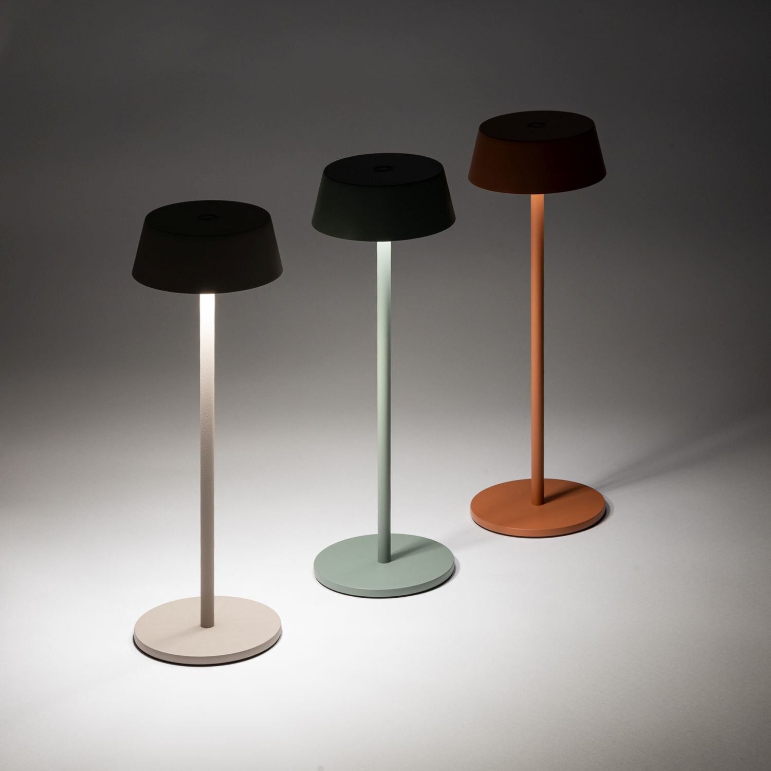 Trzy przenośne lampy zewnętrzne LED o różnych kolorach podstawy i klosza: beżowym, zielonym i pomarańczowym. Każda lampa ma prosty, cylindryczny kształt z okrągłą podstawą i kloszem.
