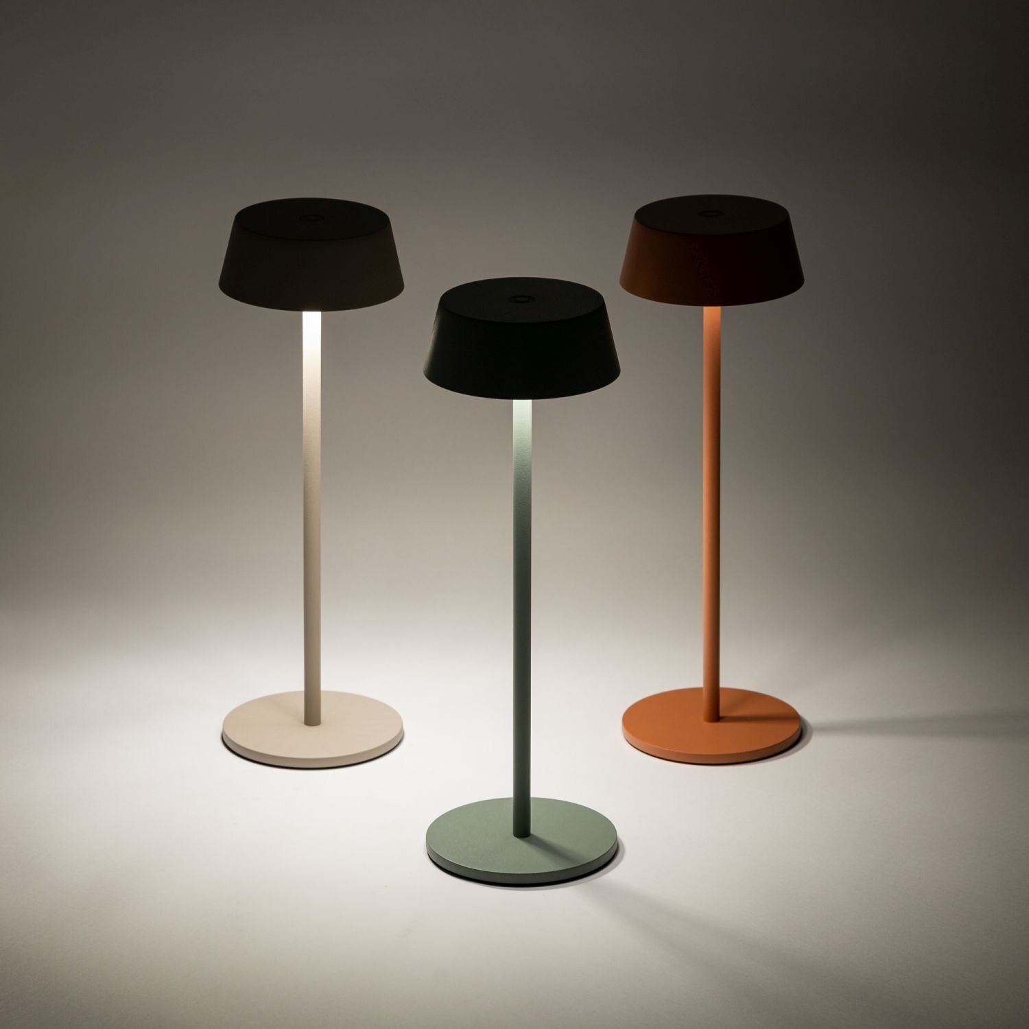 Na obrazie widoczne są trzy przenośne lampy zewnętrzne LED o prostym, nowoczesnym designie, każda w innym kolorze: beżowym, zielonym i pomarańczowym. Lampy mają okrągłe podstawy i klosze, emitując delikatne światło.