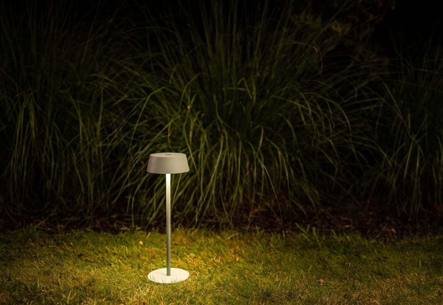 Na trawniku stoi przenośna lampa zewnętrzna LED, emitująca ciepłe światło. W tle widoczne są wysokie trawy.