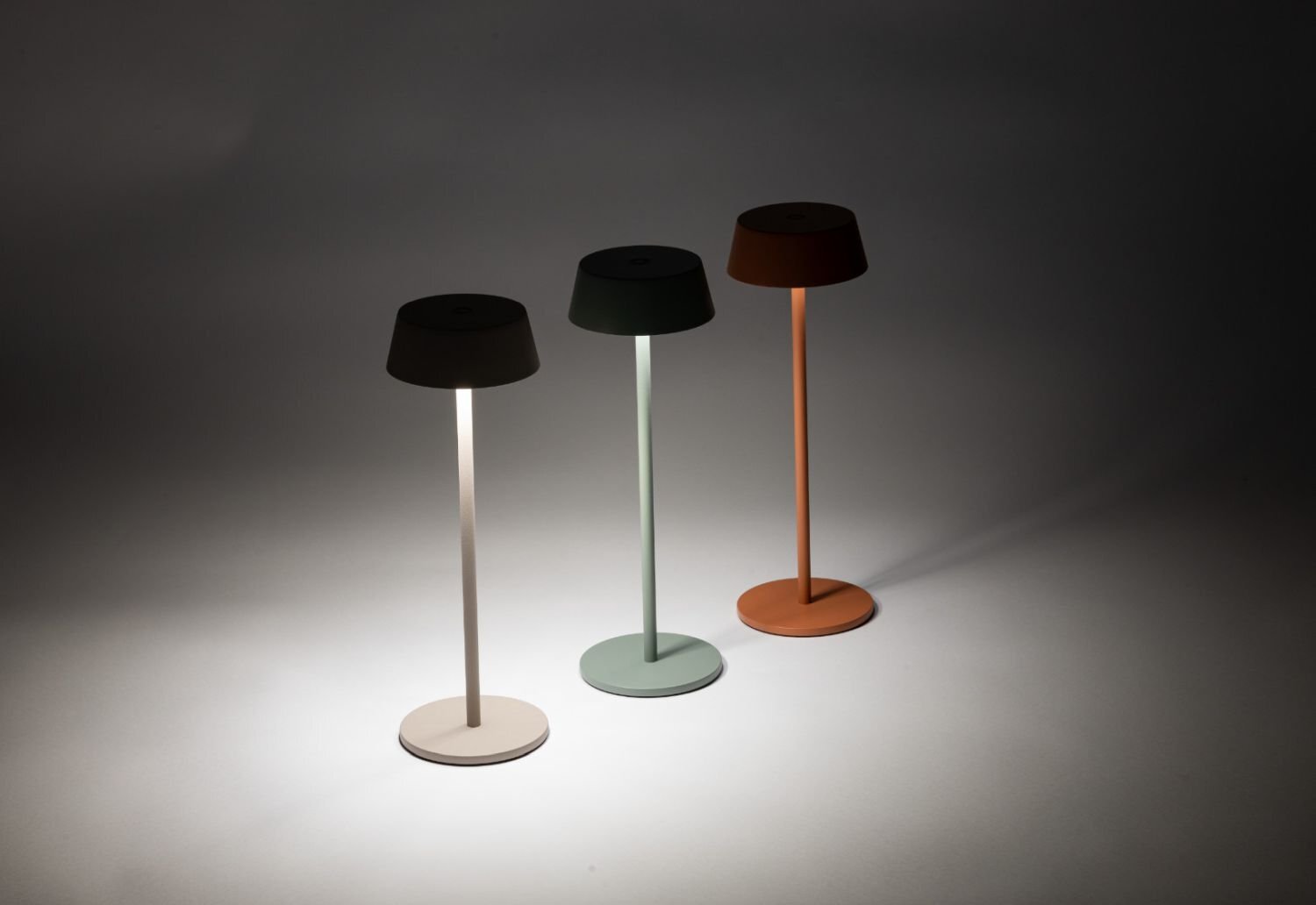 Na obrazie widoczne są trzy przenośne lampy zewnętrzne LED o prostym, nowoczesnym designie, każda w innym kolorze: białym, zielonym i pomarańczowym. Lampy stoją na płaskiej powierzchni, a jedna z nich jest włączona, emitując światło.
