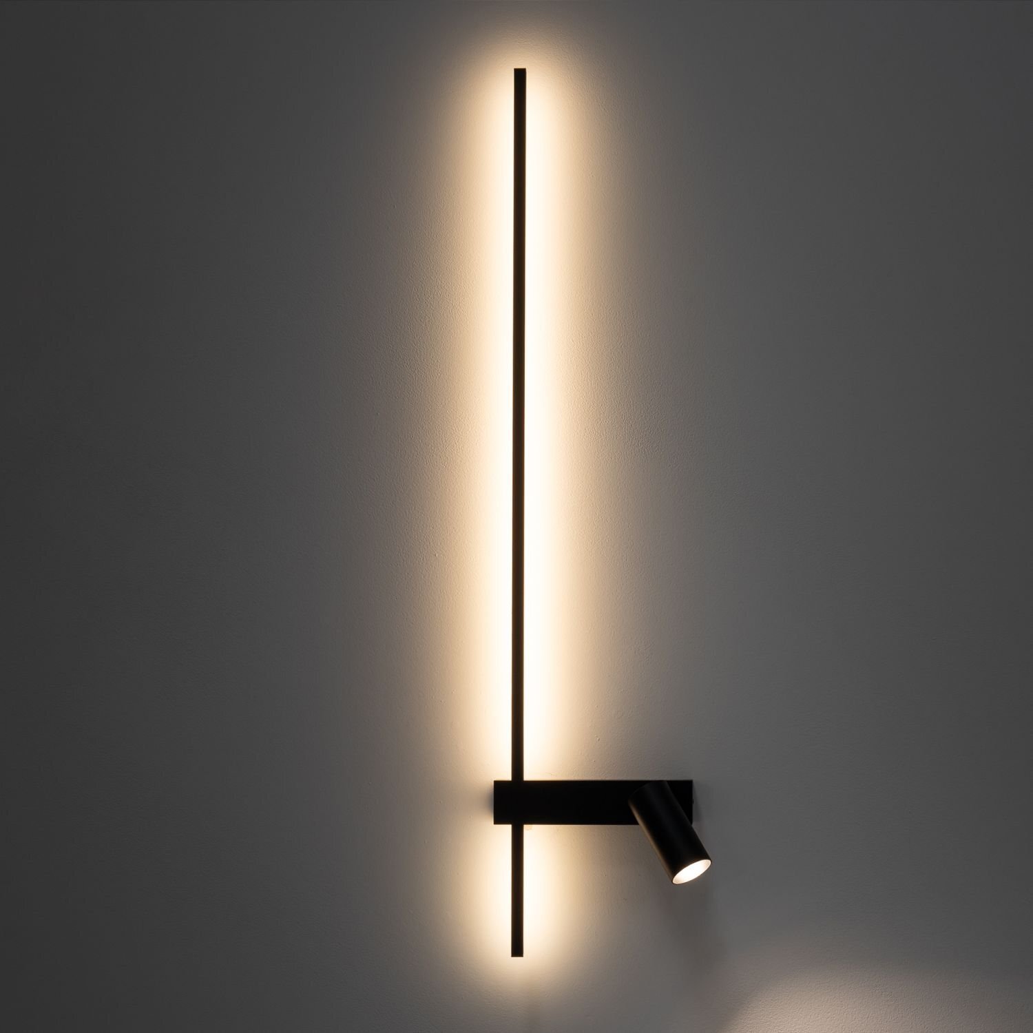 Na obrazie widoczna jest nowoczesna lampa ścienna z długim, pionowym paskiem LED emitującym ciepłe światło oraz reflektorem skierowanym w dół. Lampa zamontowana jest na gładkiej, szarej ścianie.