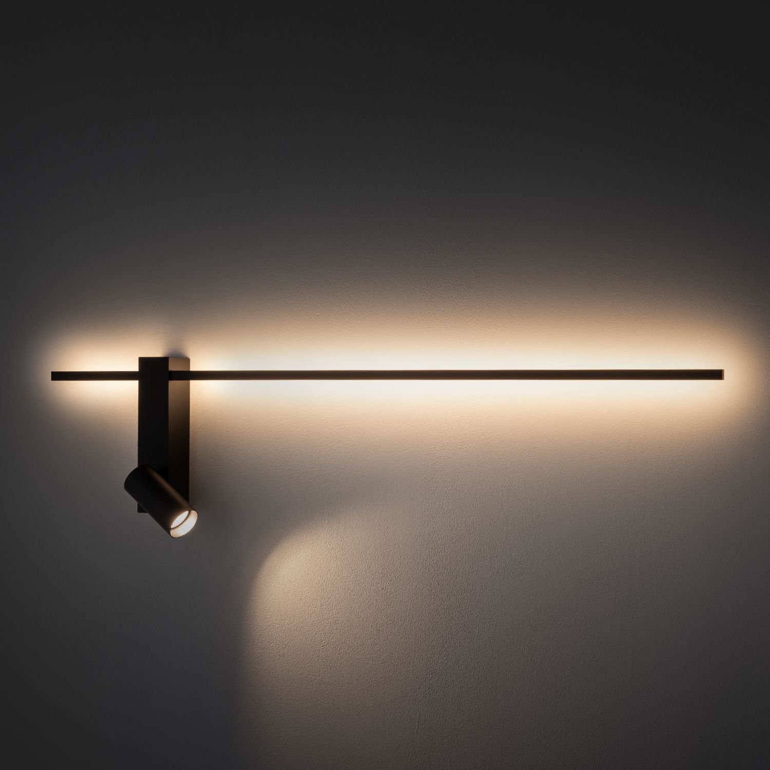 Na obrazie widoczna jest nowoczesna lampa ścienna z reflektorem, emitująca ciepłe światło na ścianę. Lampa ma prosty, minimalistyczny design z długim, poziomym elementem świetlnym.