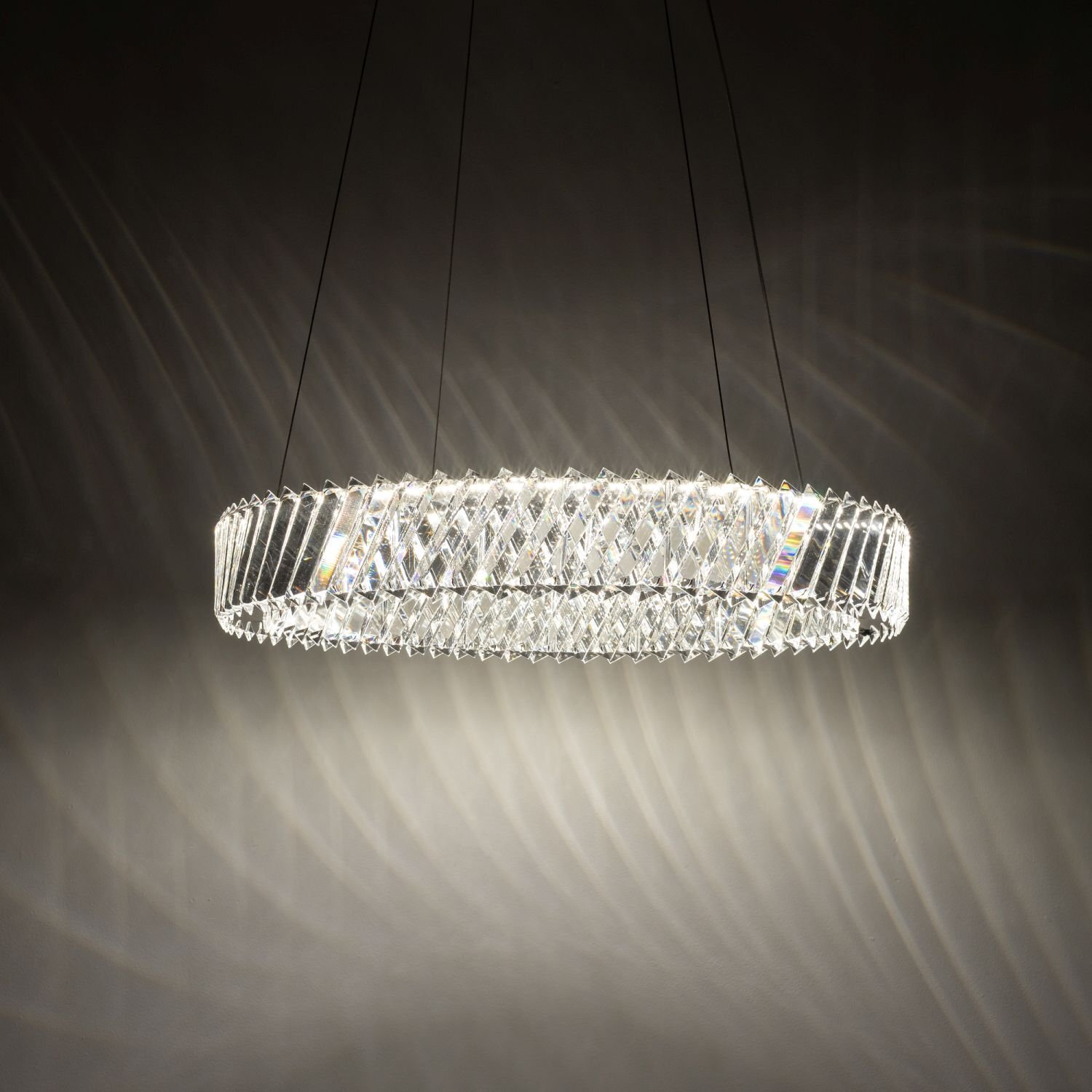 Na obrazie widoczna jest lampa wisząca o okrągłym kształcie, wykonana z przezroczystych elementów, które tworzą efekt świetlny na ścianie. Lampa jest zawieszona na cienkich przewodach i emituje jasne światło.