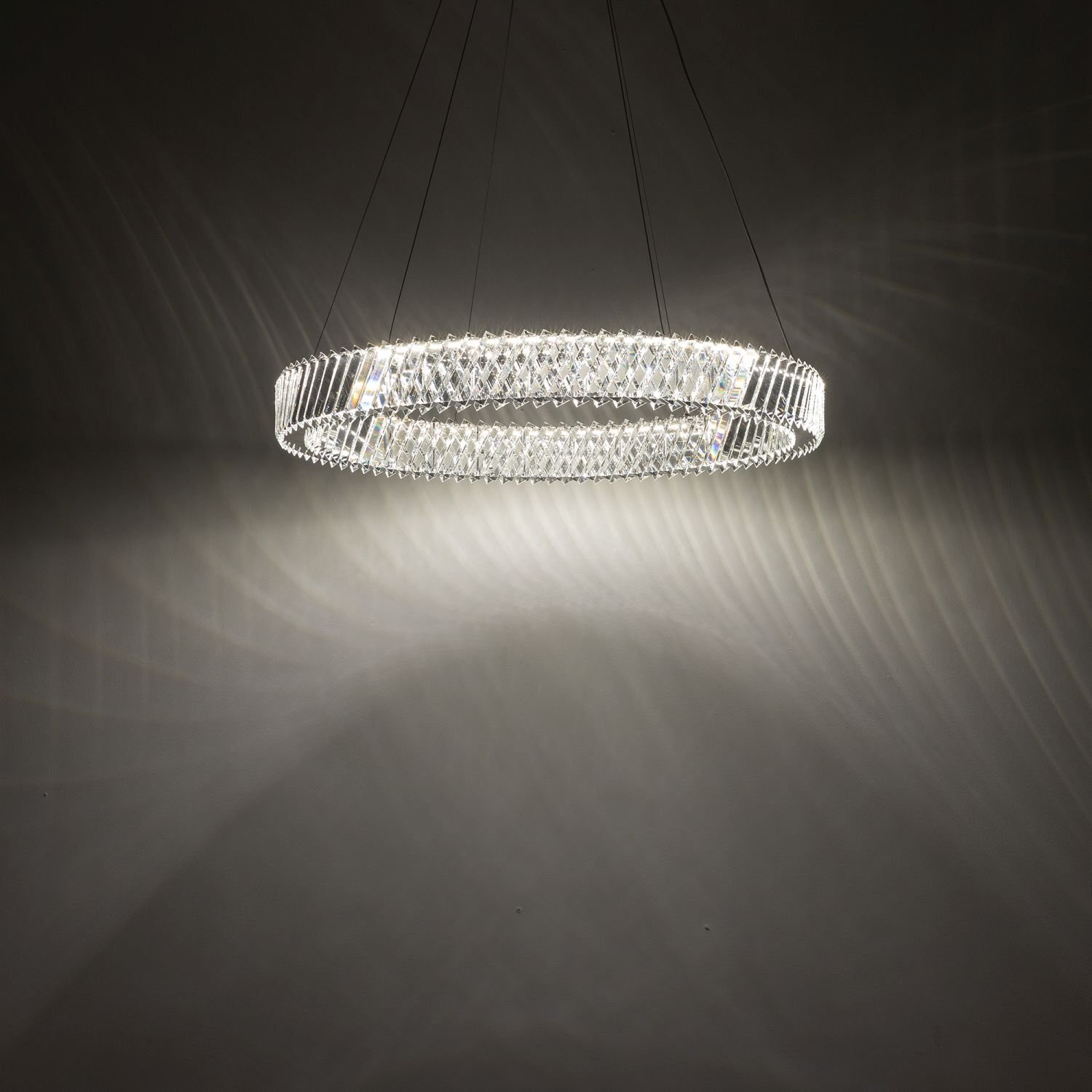 Na obrazie widoczna jest okrągła lampa wisząca LED, emitująca jasne światło, które tworzy wzory na ścianie. Lampa zawieszona jest na cienkich przewodach, a jej powierzchnia ma strukturę przypominającą kryształki.