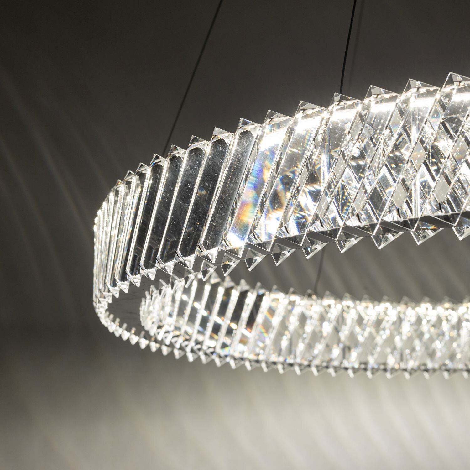 Zdjęcie przedstawia wiszącą lampę LED o nowoczesnym designie, złożoną z przezroczystych, geometrycznych elementów tworzących okrągły kształt. Światło odbija się od powierzchni, tworząc efektowne refleksy.