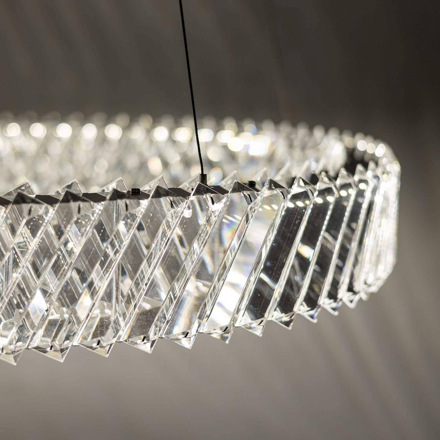 Zdjęcie przedstawia zbliżenie na wiszącą lampę LED z przezroczystymi, geometrycznymi elementami tworzącymi okrągły kształt. Lampa jest zawieszona na cienkich przewodach, a światło odbija się od jej powierzchni, tworząc efektowne refleksy.