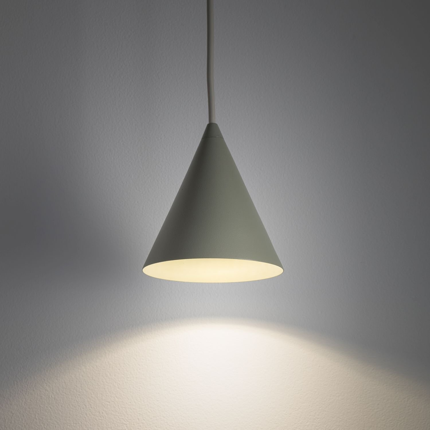 Na obrazie widoczna jest wisząca lampa o stożkowym kształcie, emitująca ciepłe światło na ścianę. Lampa jest zawieszona na cienkim przewodzie.