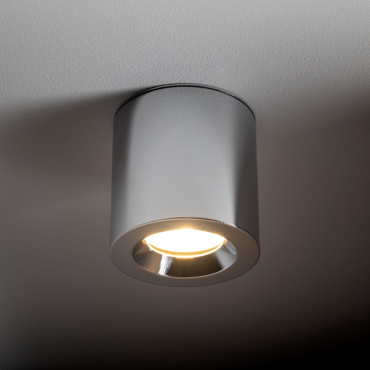Na obrazie widoczna jest elegancka, cylindryczna lampa sufitowa emitująca ciepłe światło. Lampa jest zamontowana na gładkim suficie.