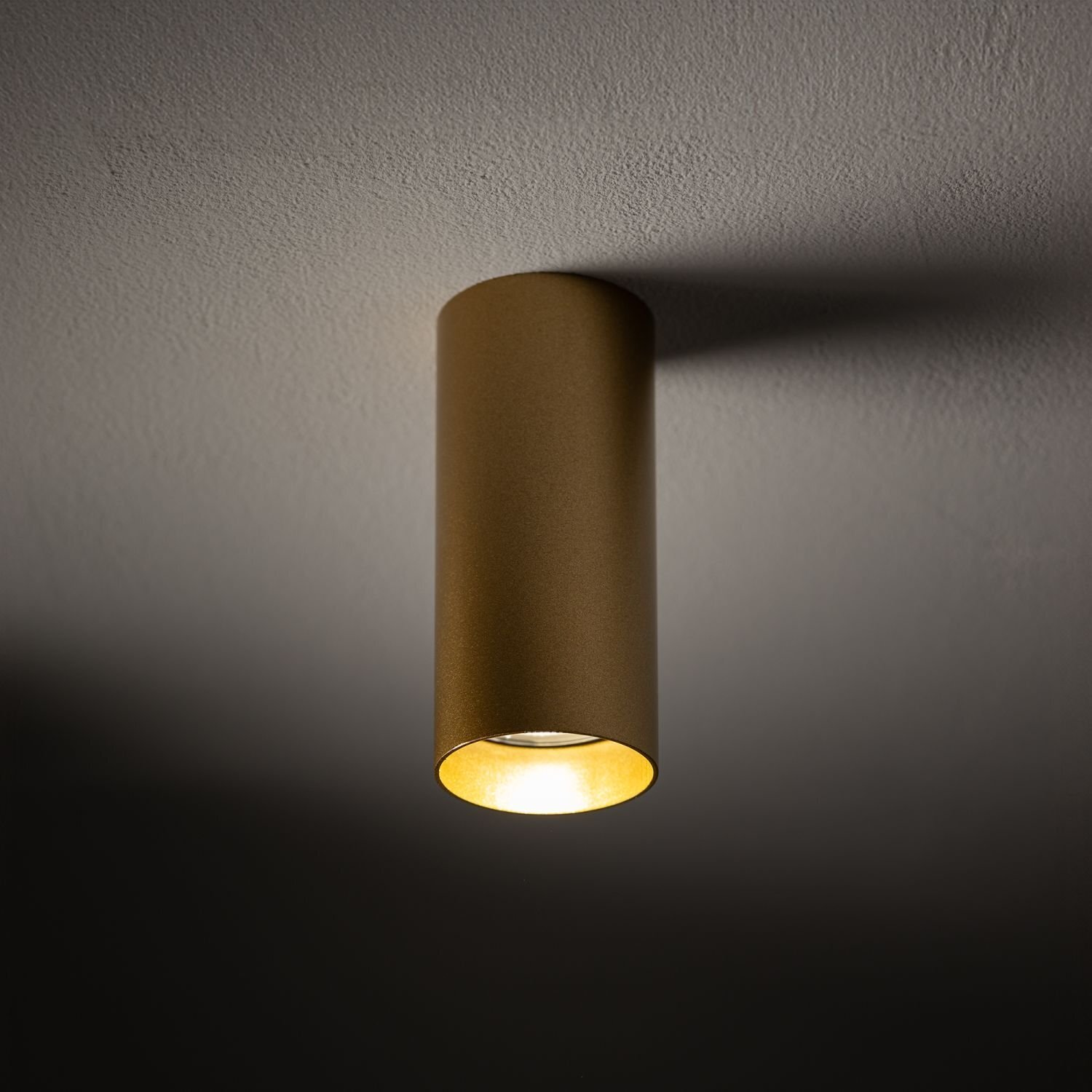 Na obrazie widoczna jest cylindryczna lampa sufitowa w kolorze złotym, emitująca ciepłe światło. Lampa jest zamontowana na jasnym suficie.
