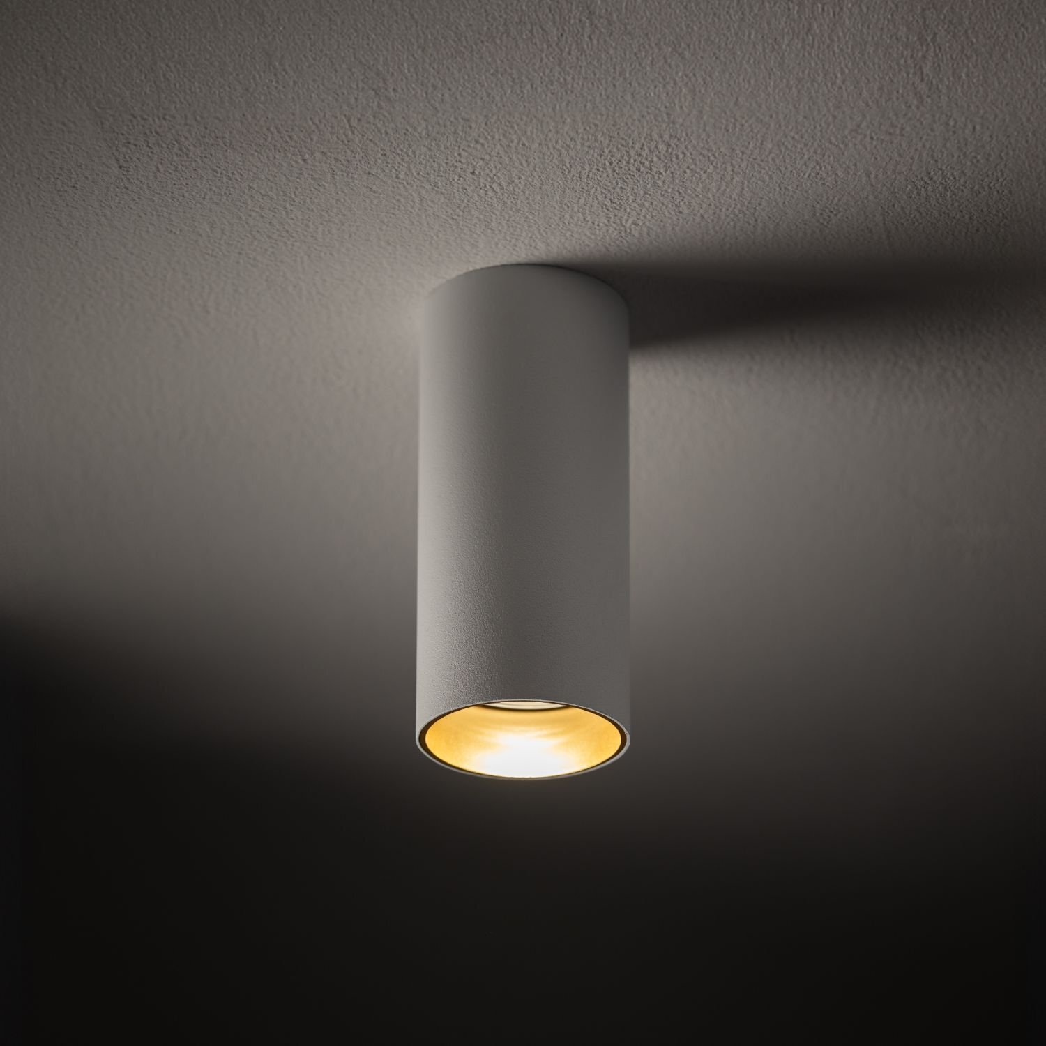 Cylindryczna lampa natynkowa w kolorze szarym zamontowana na suficie, emitująca ciepłe światło.