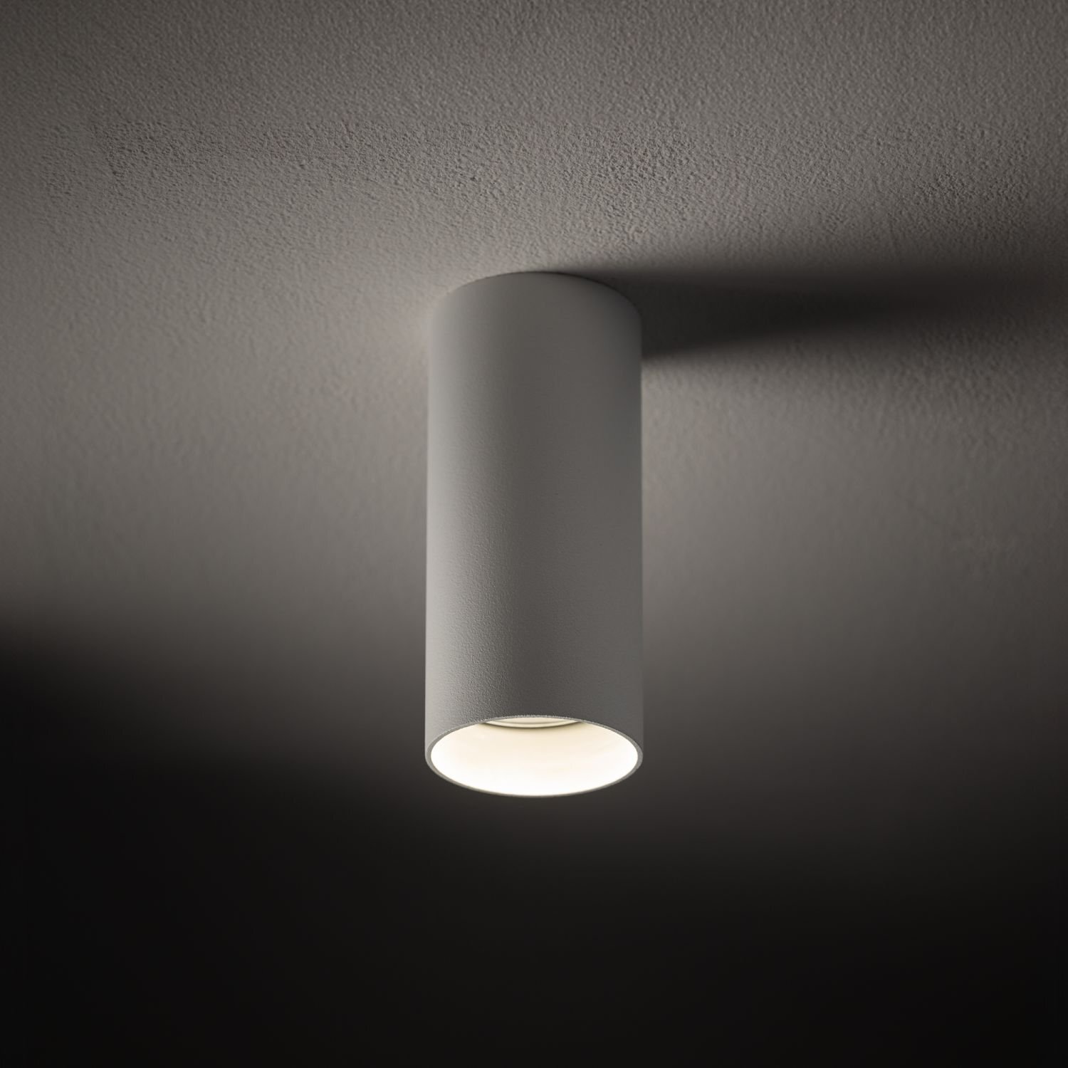 Cylindryczna lampa natynkowa w kolorze szarym zamontowana na suficie, emitująca jasne światło. Powierzchnia sufitu jest gładka i jednolita.