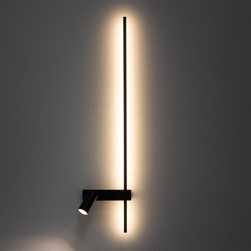 Na obrazie widoczna jest nowoczesna lampa ścienna z długim, pionowym elementem emitującym ciepłe światło oraz reflektorem skierowanym w dół. Lampa zamontowana jest na gładkiej, ciemnej ścianie.