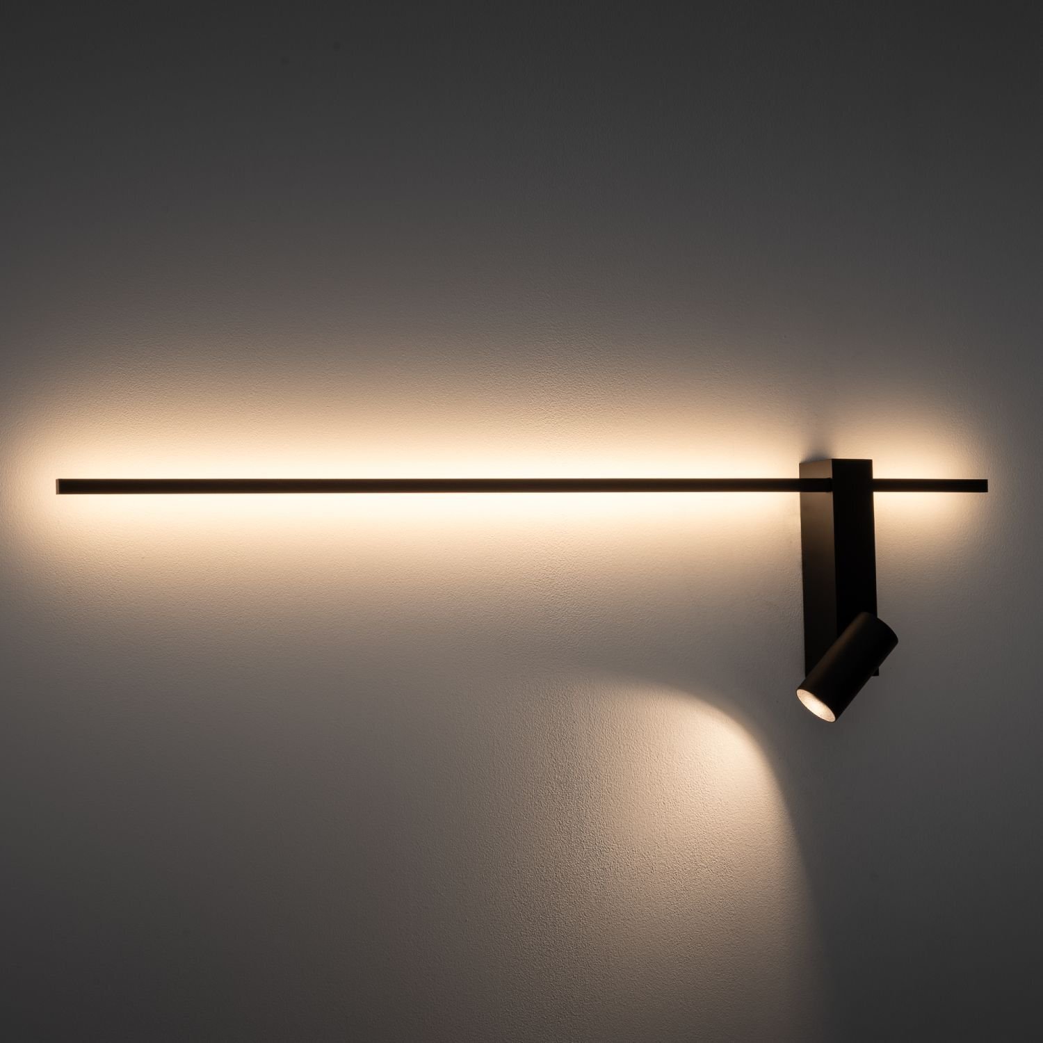 Na obrazie widoczna jest nowoczesna lampa ścienna z reflektorem, emitująca ciepłe światło na ścianę. Lampa ma prosty, minimalistyczny design z długim, poziomym elementem i skierowanym w dół reflektorem.