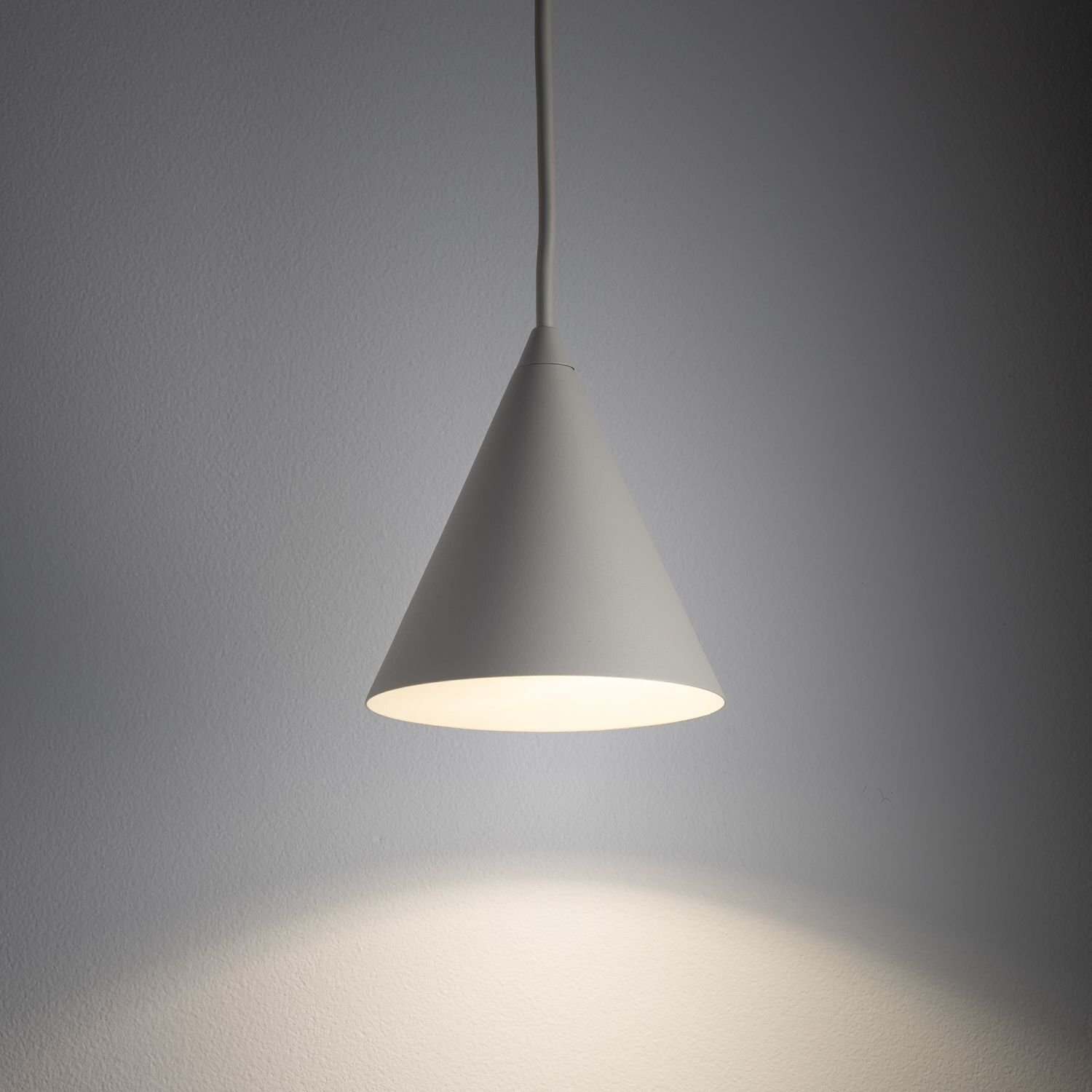 Na obrazie widoczna jest wisząca lampa w kształcie stożka, emitująca światło na ścianę. Lampa ma jednolity, jasny kolor.