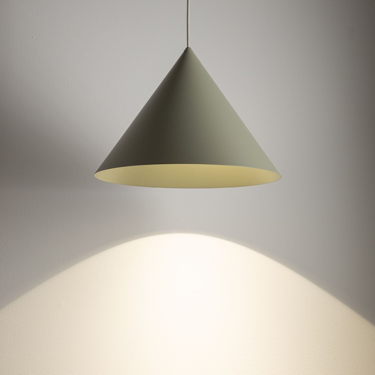 Na obrazie widoczna jest wisząca lampa w kształcie stożka, emitująca światło na ścianę. Lampa ma jednolity, jasny kolor i jest zawieszona na cienkim przewodzie.