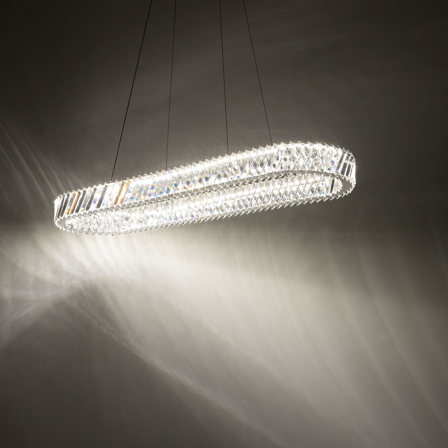 Na obrazie widoczna jest nowoczesna lampa wisząca LED o owalnym kształcie, emitująca jasne światło. Lampa zawieszona jest na cienkich przewodach, a jej powierzchnia ma strukturę przypominającą kryształki, które rozpraszają światło na ścianie.