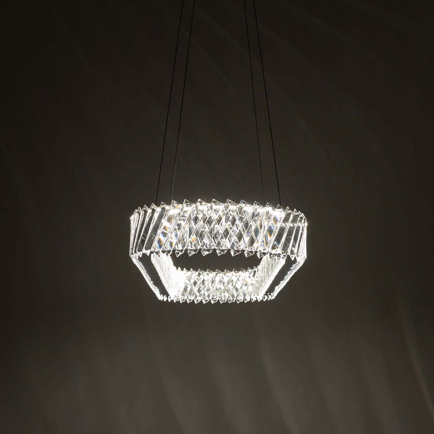 Na obrazie widoczna jest nowoczesna lampa wisząca LED o geometrycznym kształcie, emitująca jasne światło. Lampa zawieszona jest na cienkich przewodach, a jej konstrukcja składa się z przezroczystych elementów tworzących wzór przypominający kryształ.