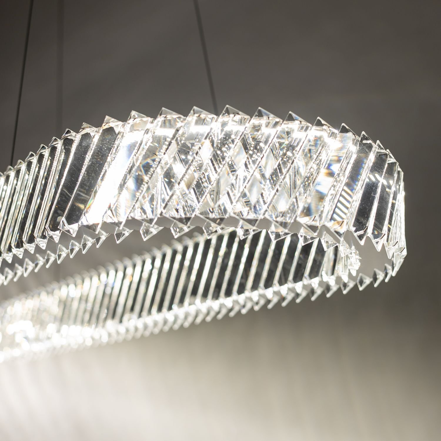 Obraz przedstawia nowoczesną lampę wiszącą LED z przezroczystymi, geometrycznymi elementami, które odbijają światło. Lampa emituje jasne, białe światło.