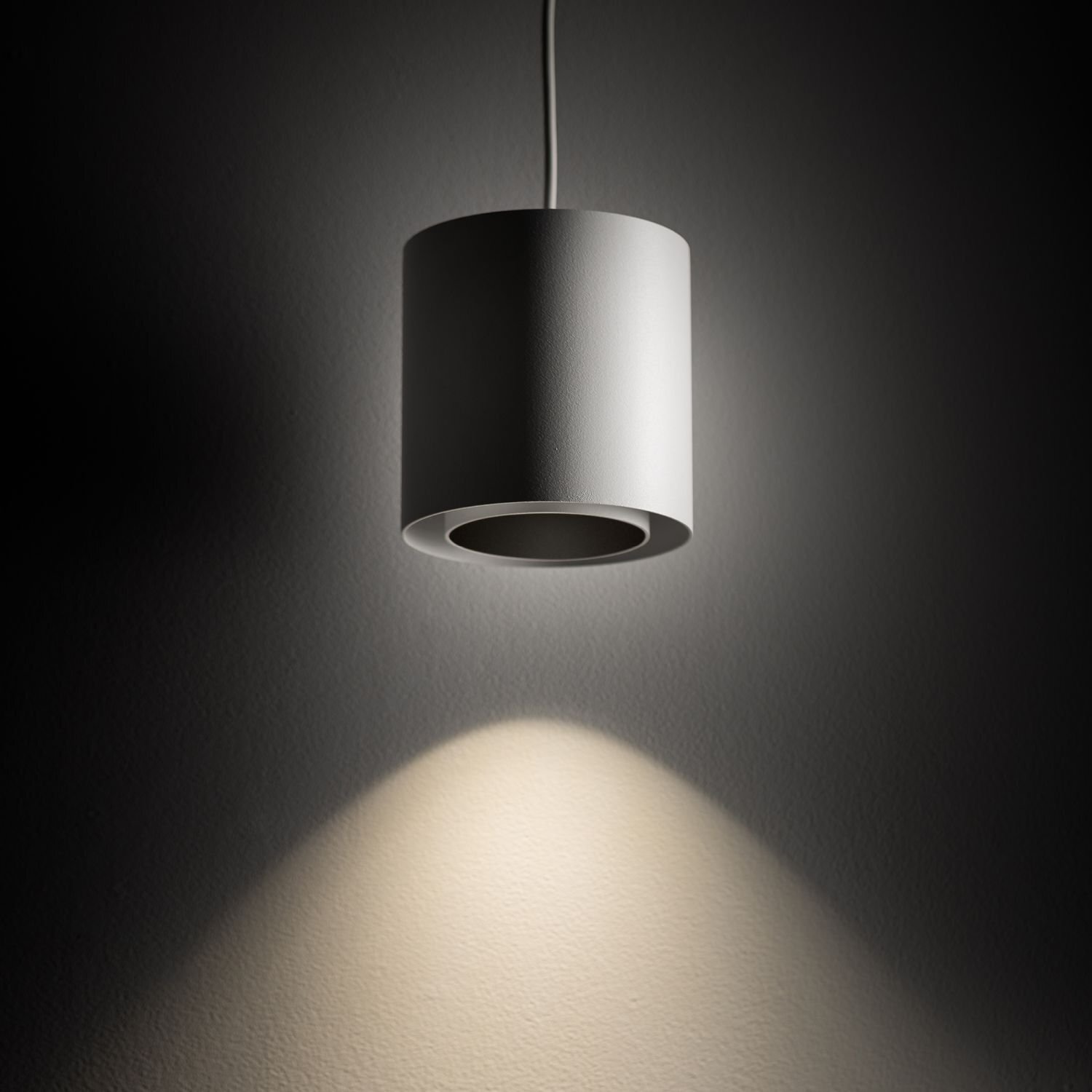 Na obrazie widoczna jest nowoczesna, cylindryczna lampa wisząca w kolorze białym, emitująca światło skierowane w dół. Światło tworzy jasną plamę na ciemnym tle ściany.