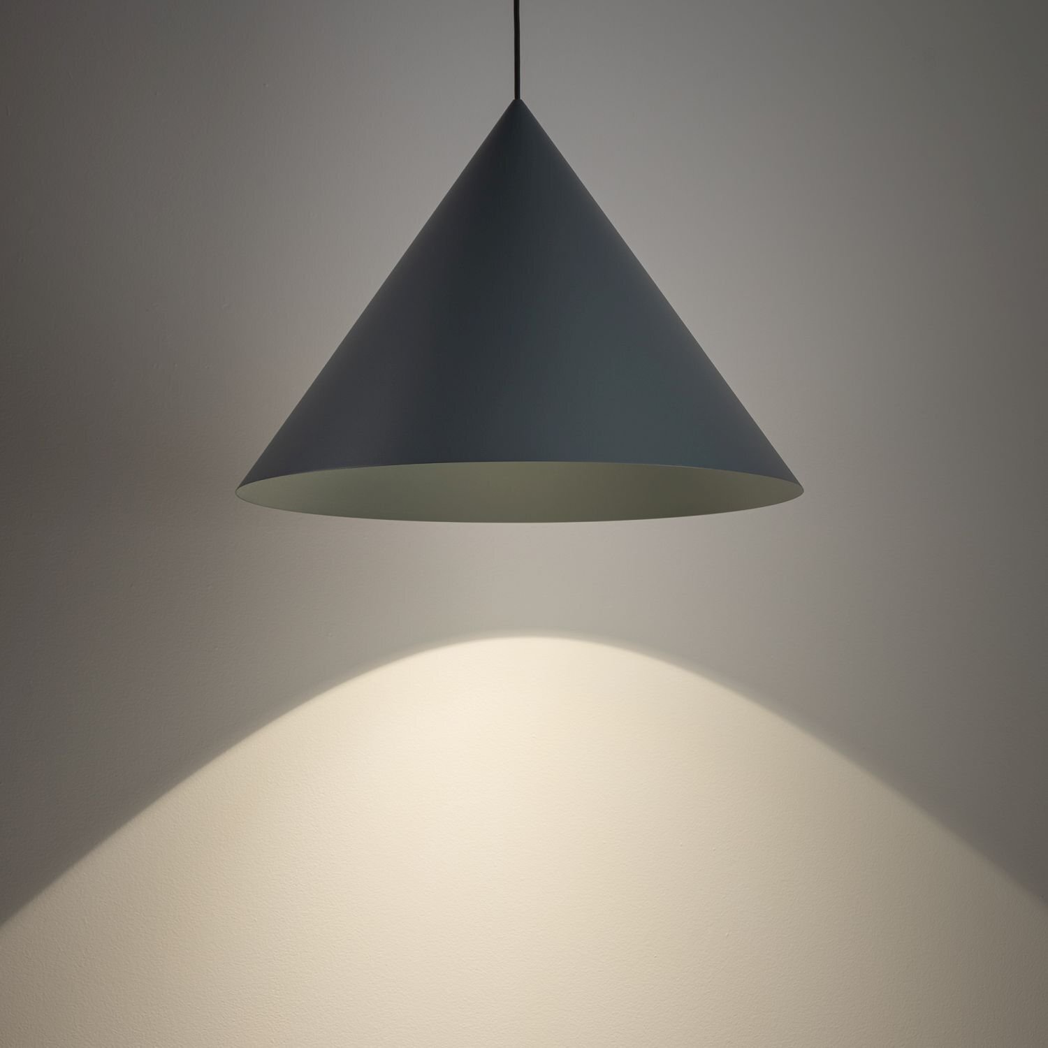 Na obrazie widoczna jest nowoczesna lampa wisząca o stożkowym kształcie, emitująca światło na ścianę. Lampa jest w ciemnym kolorze, a światło tworzy jasną plamę na powierzchni poniżej.