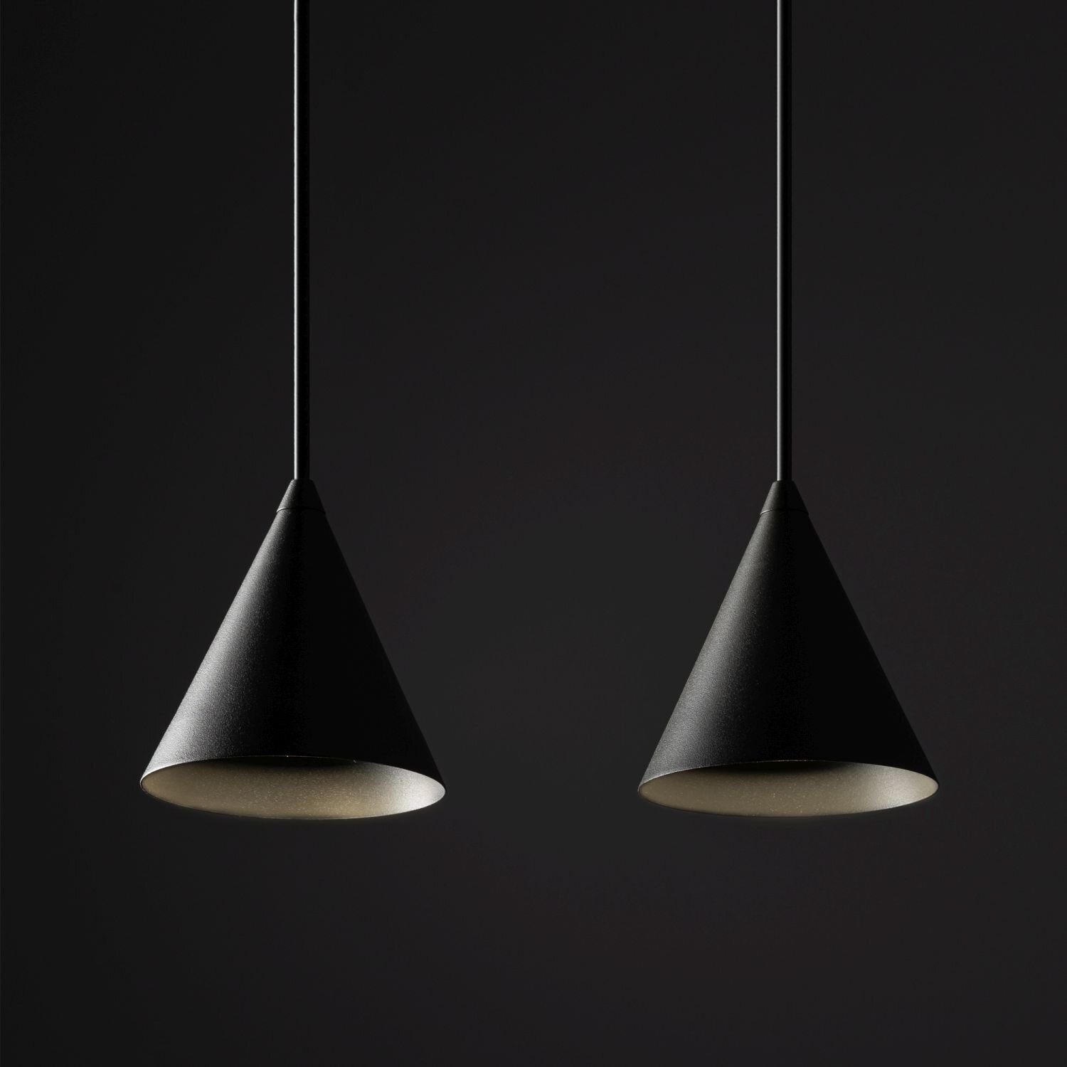Na obrazie widoczne są dwie czarne, stożkowe lampy wiszące na ciemnym tle. Lampy mają gładką powierzchnię i emitują światło skierowane w dół.