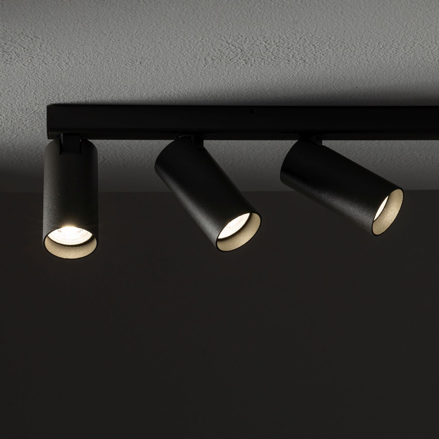 Czarna lampa sufitowa z trzema cylindrycznymi reflektorami skierowanymi w dół, zamontowana na prostokątnej listwie. Światło jest włączone, oświetlając ciemne tło.
